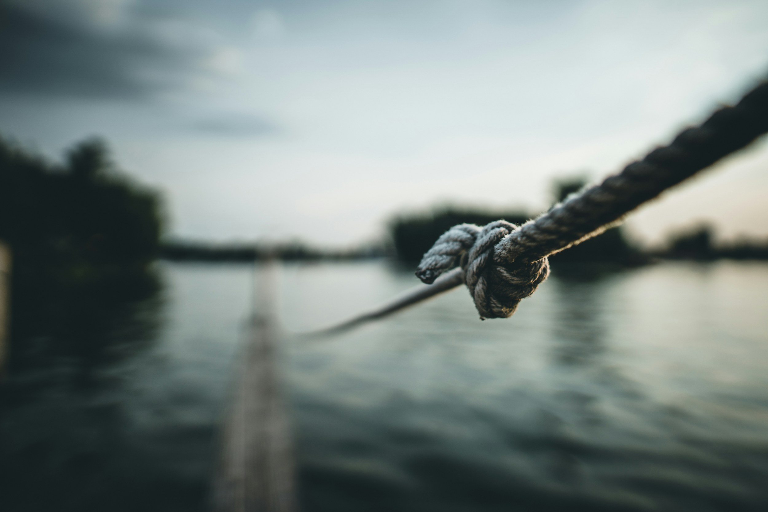 Close-up of a weathered rope tied in a knot, extending over water with a blurry shoreline and trees in the background, under a cloudy sky.