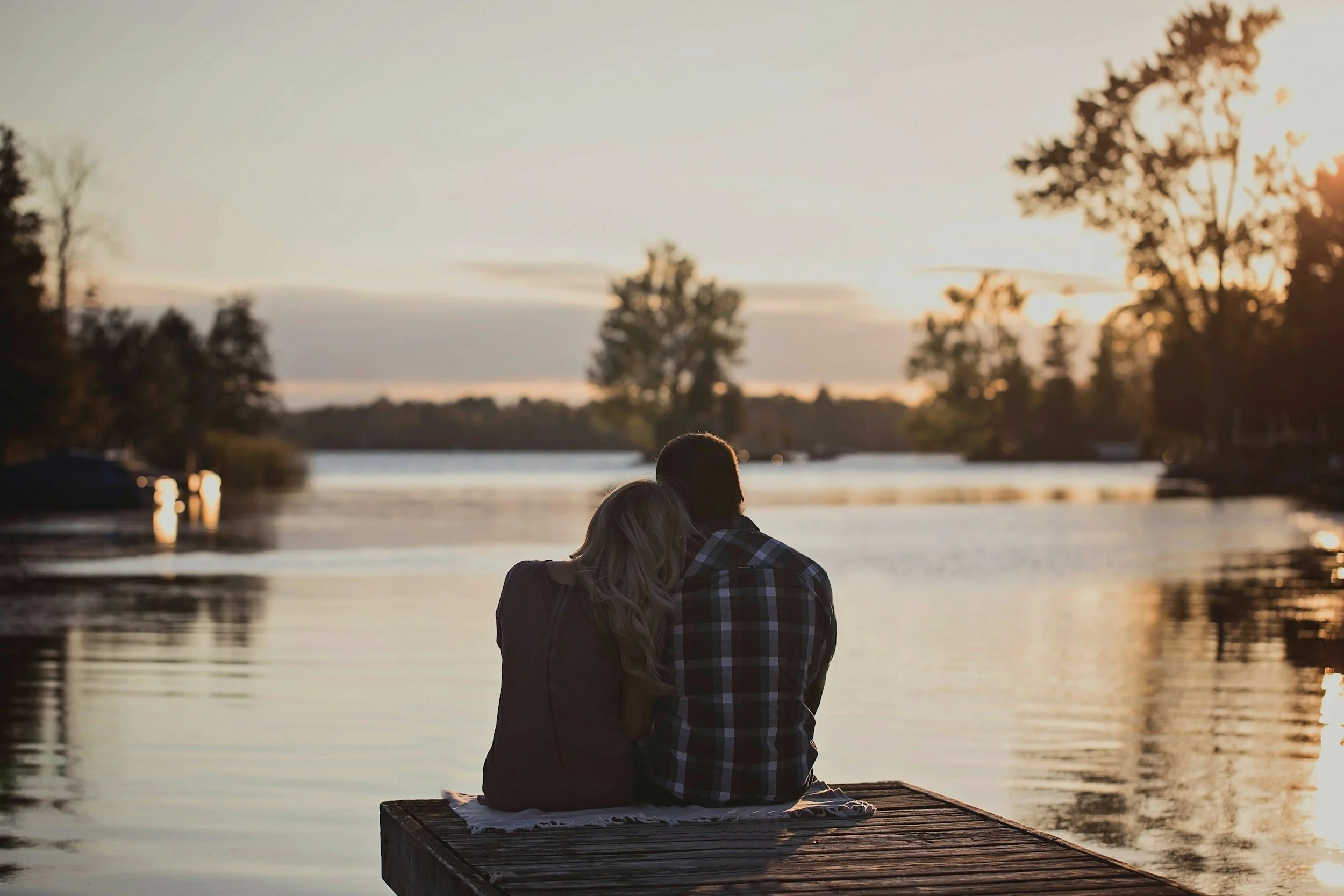 A couple sitting on a dock by a lake at sunset, embracing each other and watching the water.