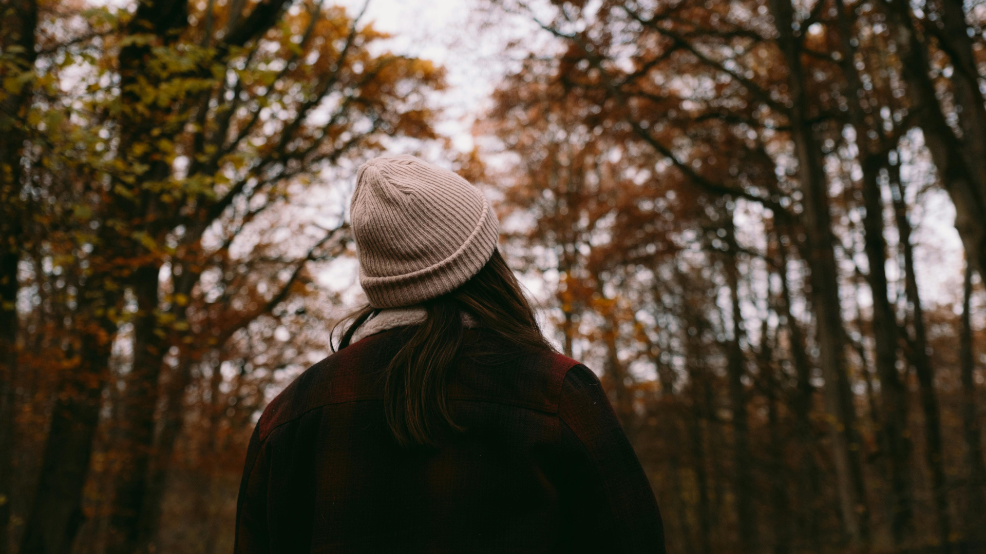 A person with long dark hair wearing a pink knit beanie and a dark jacket standing in a forest during fall with orange and yellow leaves.