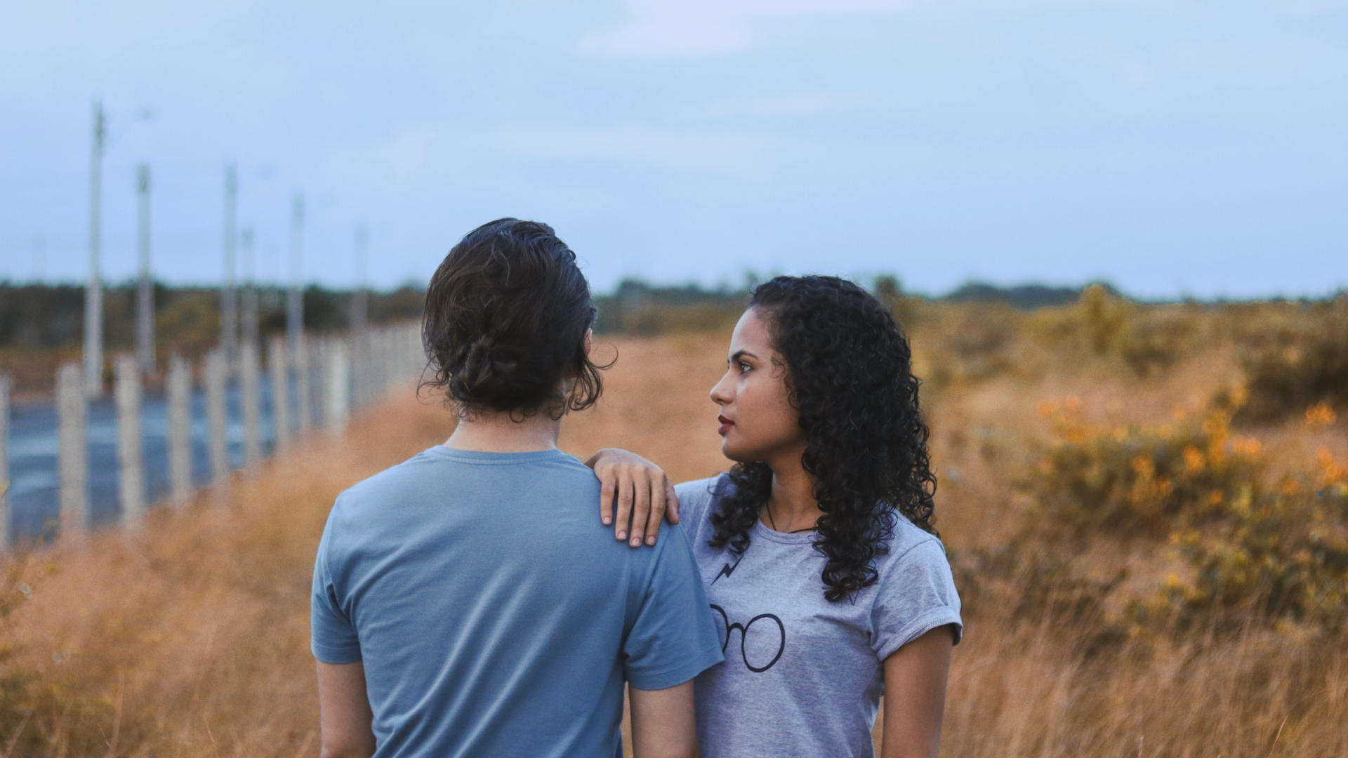 A young woman with curly dark hair and a young man with short brown hair stand closely together outdoors, with the woman resting her arm on the man's shoulder, facing each other with serious expressions. They are in a field with dry grass and a road with utility poles in the background, under a cloudy sky.