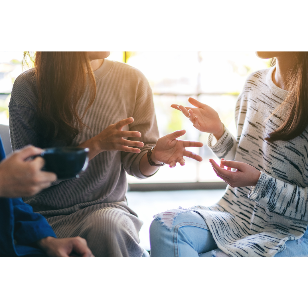 Three women talking to each other, with one holding a smartphone, indoors with sunlight in the background.