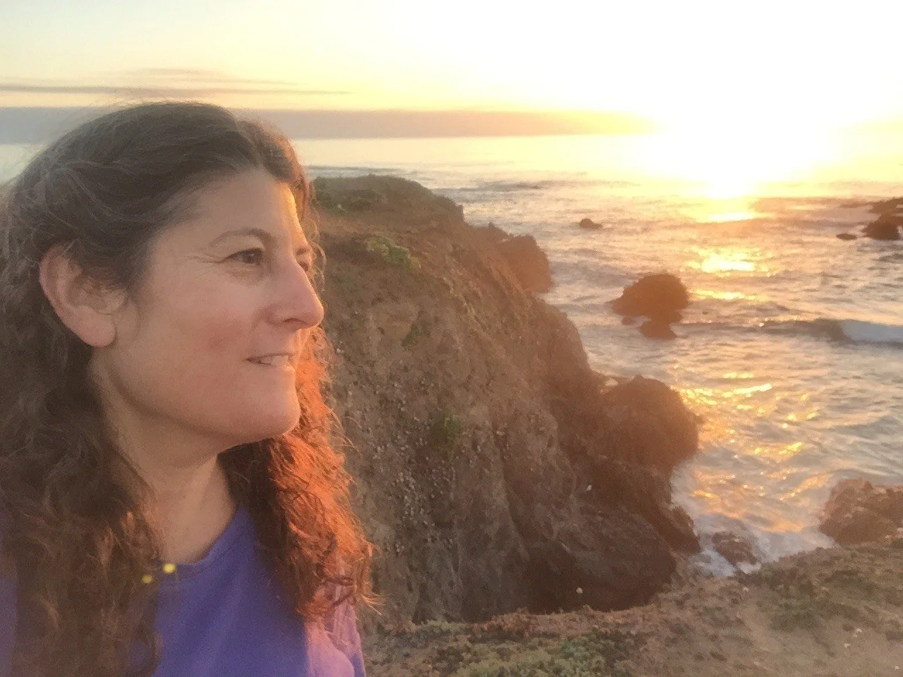 A woman with curly hair and a purple shirt gazes at the sunset over rocky coastline with ocean waves.