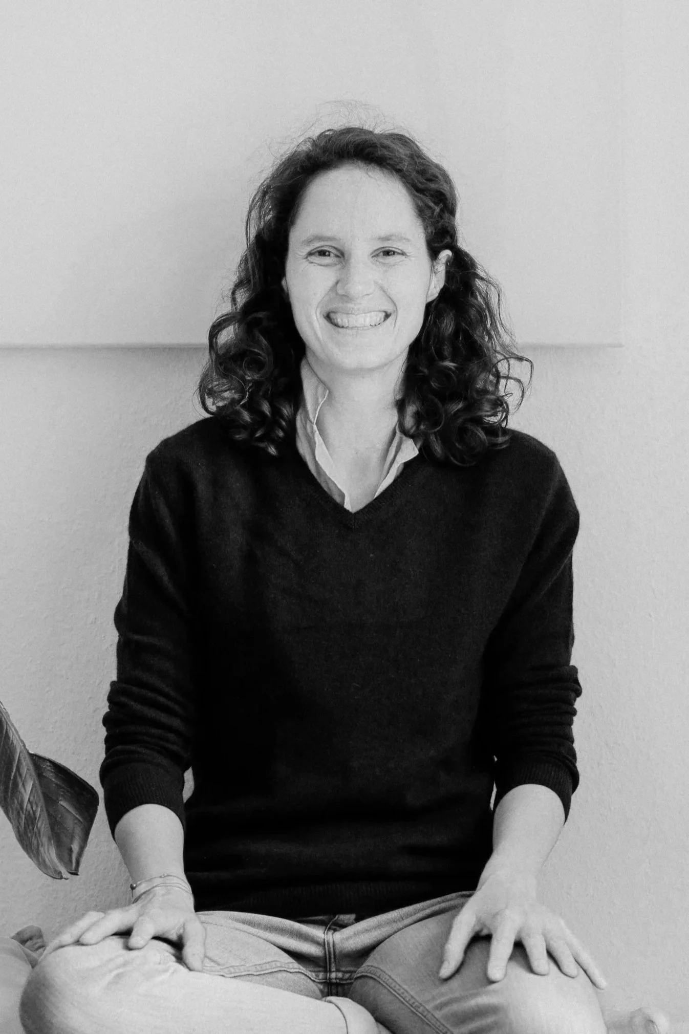Schwarz-Weiß-Foto einer lachenden Frau mit lockigem Haar, sitzend mit gekreuzten Beinen vor einer hellen Wand.