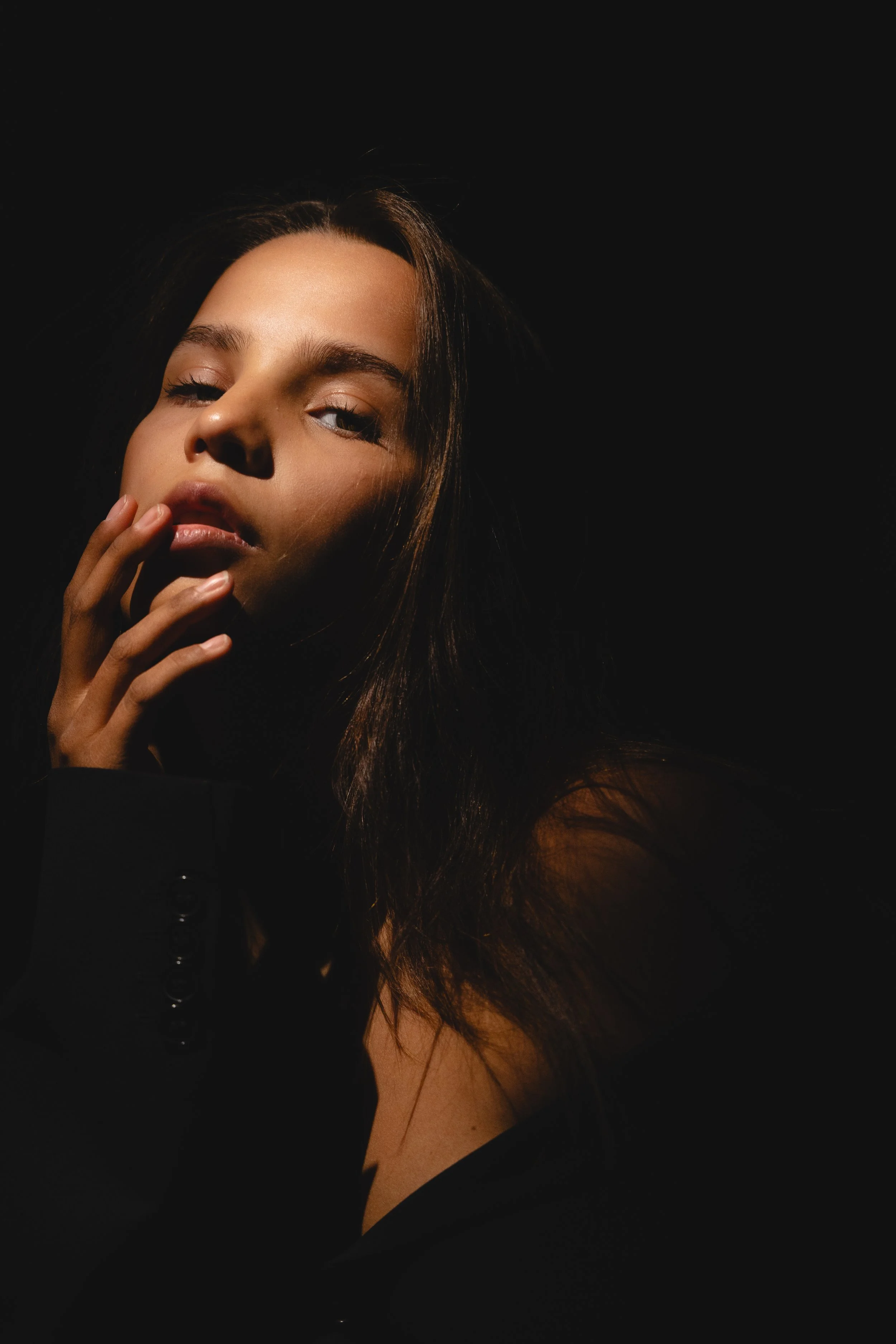 A woman with long, dark hair posing with her hand near her face, against a black background with dramatic lighting.