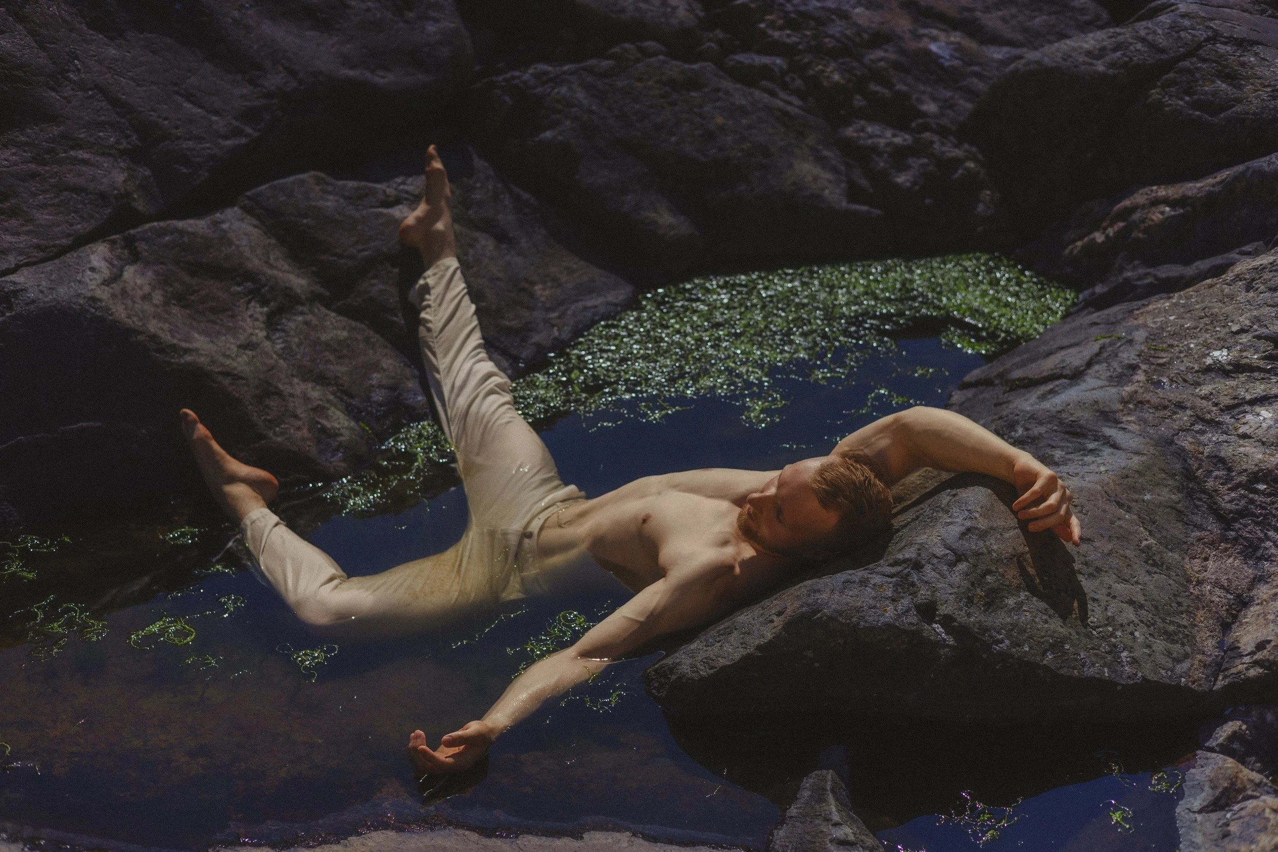 A shirtless man lying on his side in a shallow rocky pond with green algae, resting his head on a large rock and extending one arm along the water's surface.