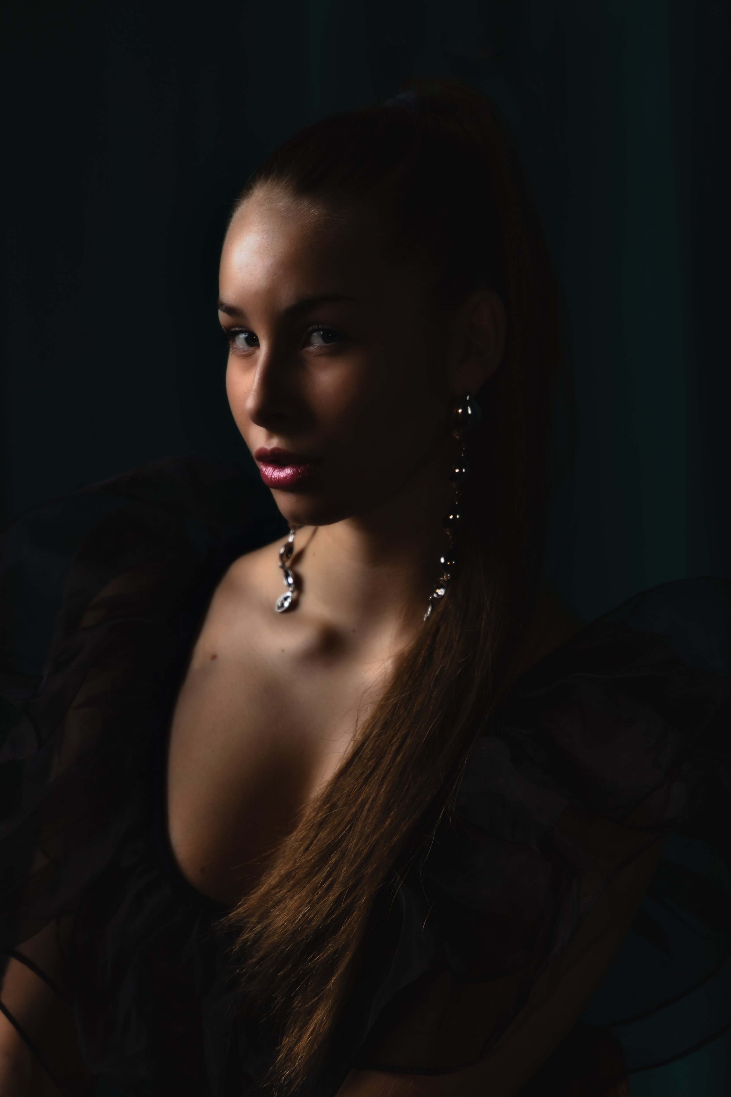 Portrait of a woman with long brown hair wearing earrings and a black, sheer, ruffled top, looking slightly to the side with a dark background.