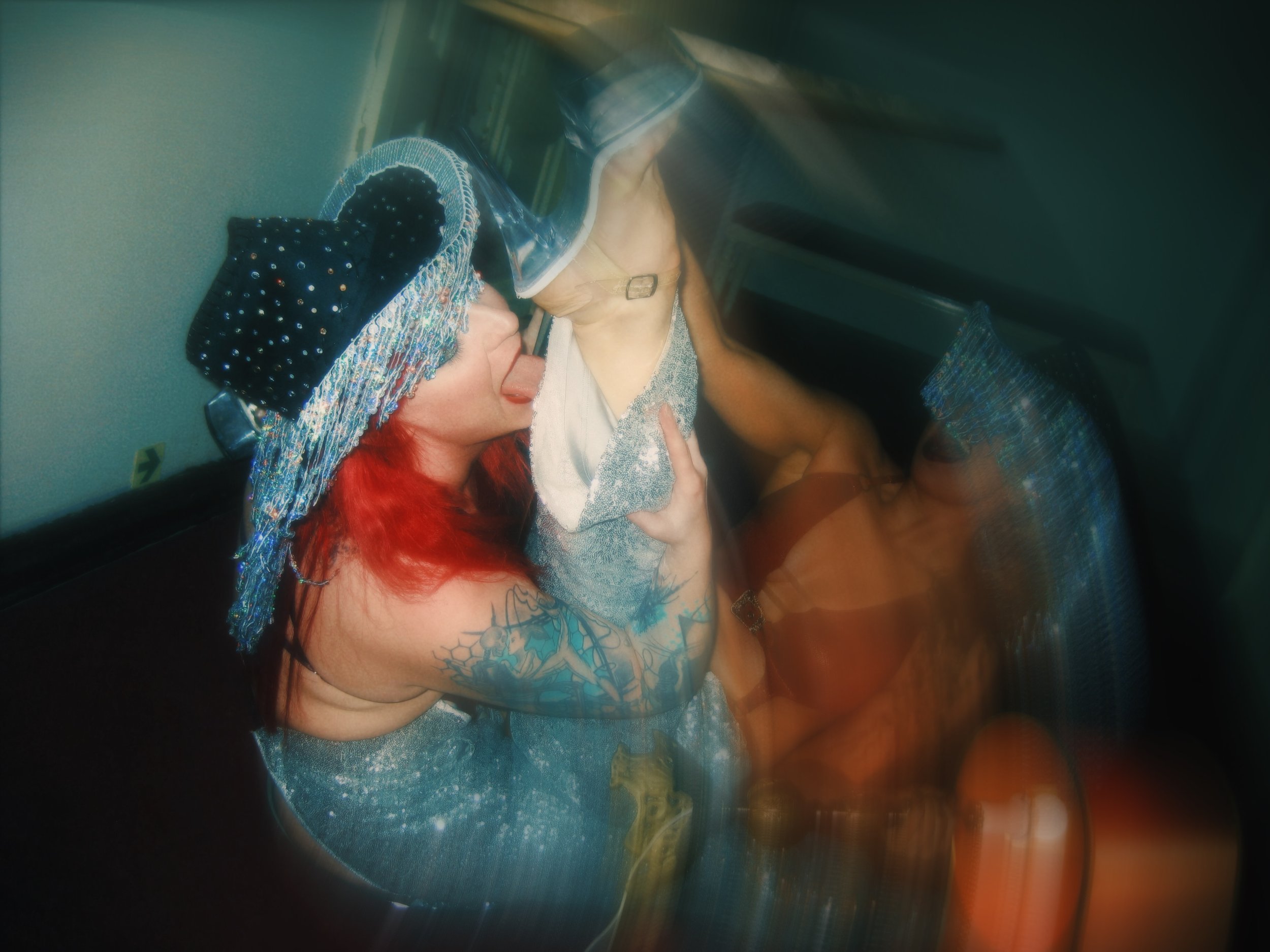 Woman with red hair and tattoos drinking from a large plastic cup, wearing a sparkly hat, sitting with her knees up.