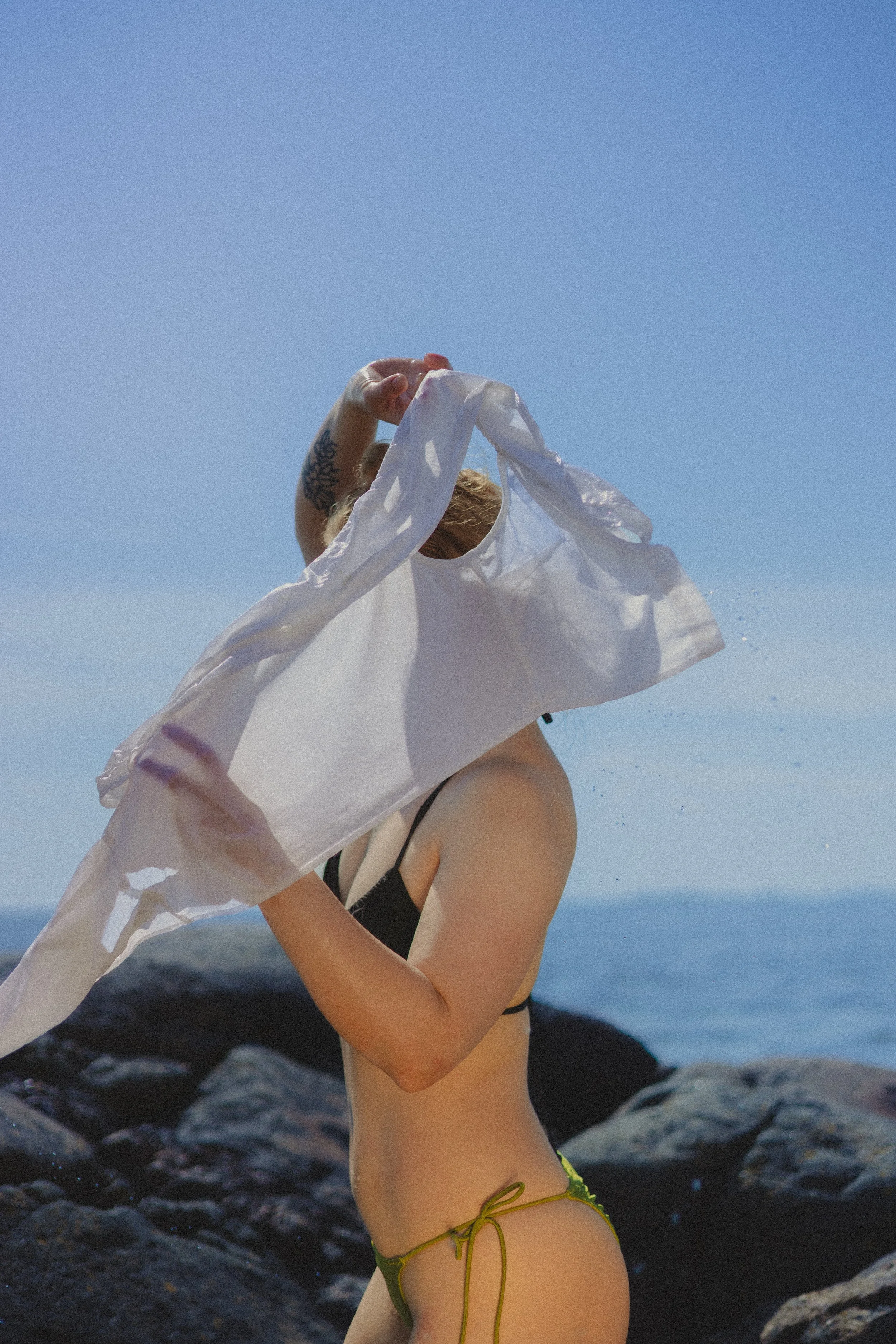 Person at the beach in a bikini, holding a white shirt over their head, with rocks and the ocean in the background.