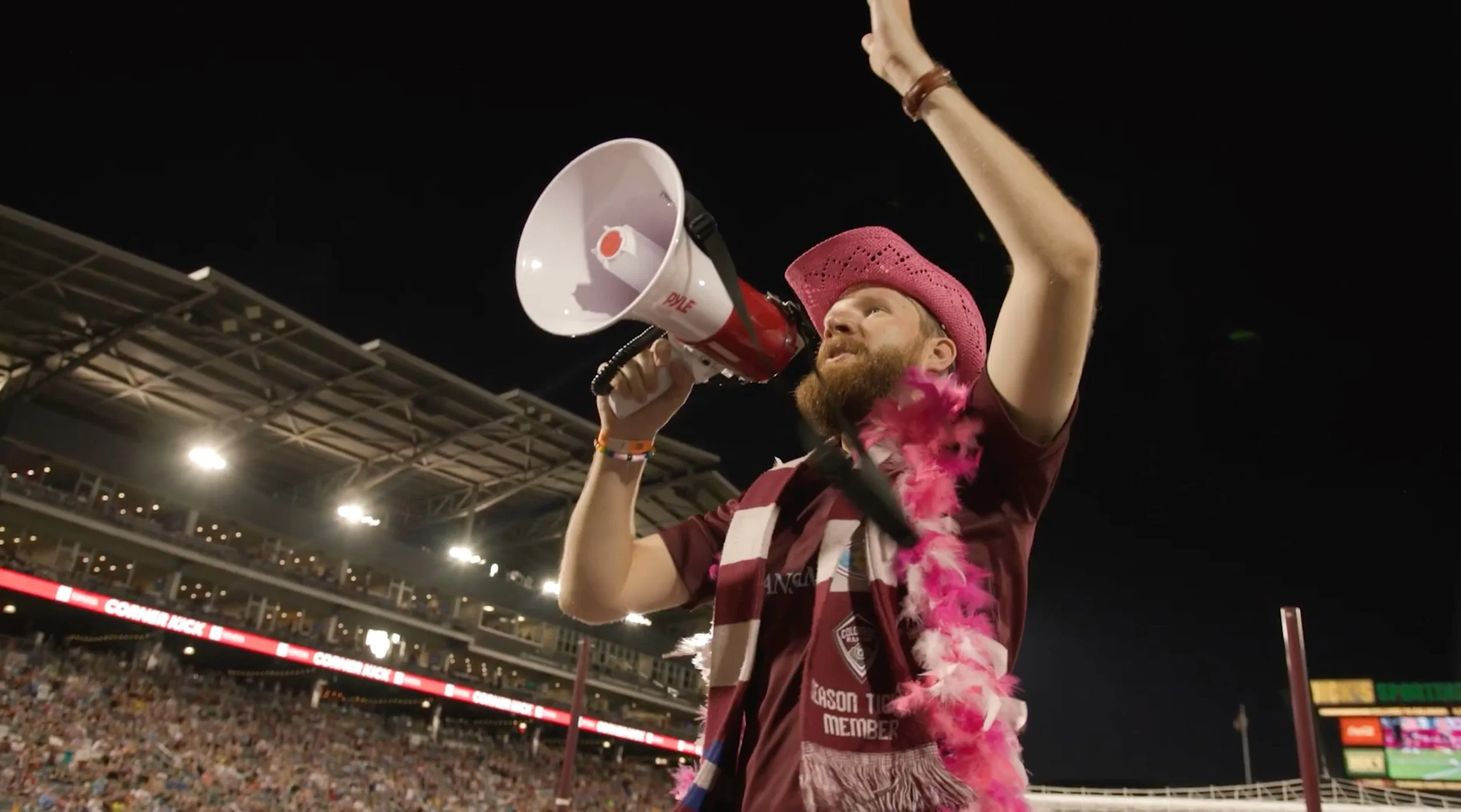 A man wearing a pink cowboy hat and pink feather boa speaking into a megaphone at a stadium during a night event, with a crowd in the background.