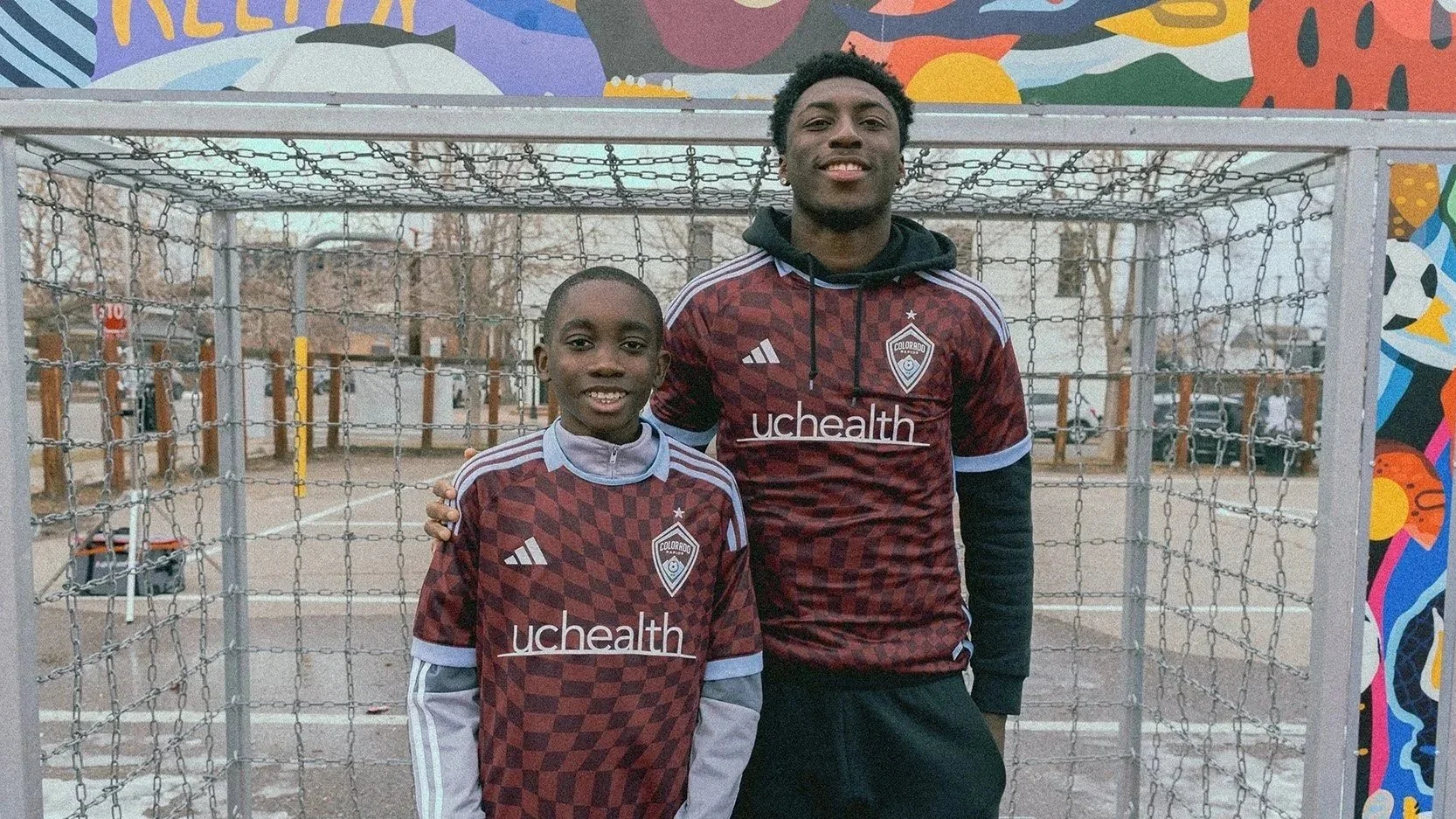 Two boys stand together on a basketball court. They are wearing matching maroon and black sports jerseys with a crest and 'uchealth' logo. A colorful mural is visible in the background.