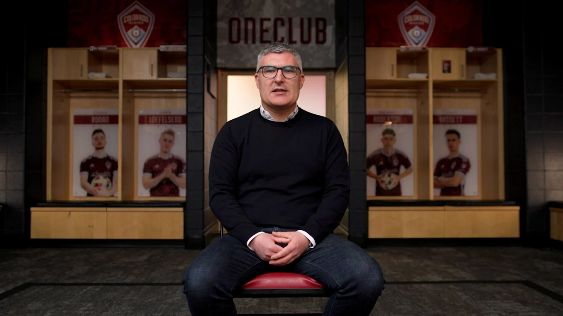 A man with gray hair and glasses wearing a black sweater and jeans is sitting on a red chair in a locker room. Behind him are wooden lockers, and posters of soccer players in maroon jerseys, holding soccer balls, are on the walls. 