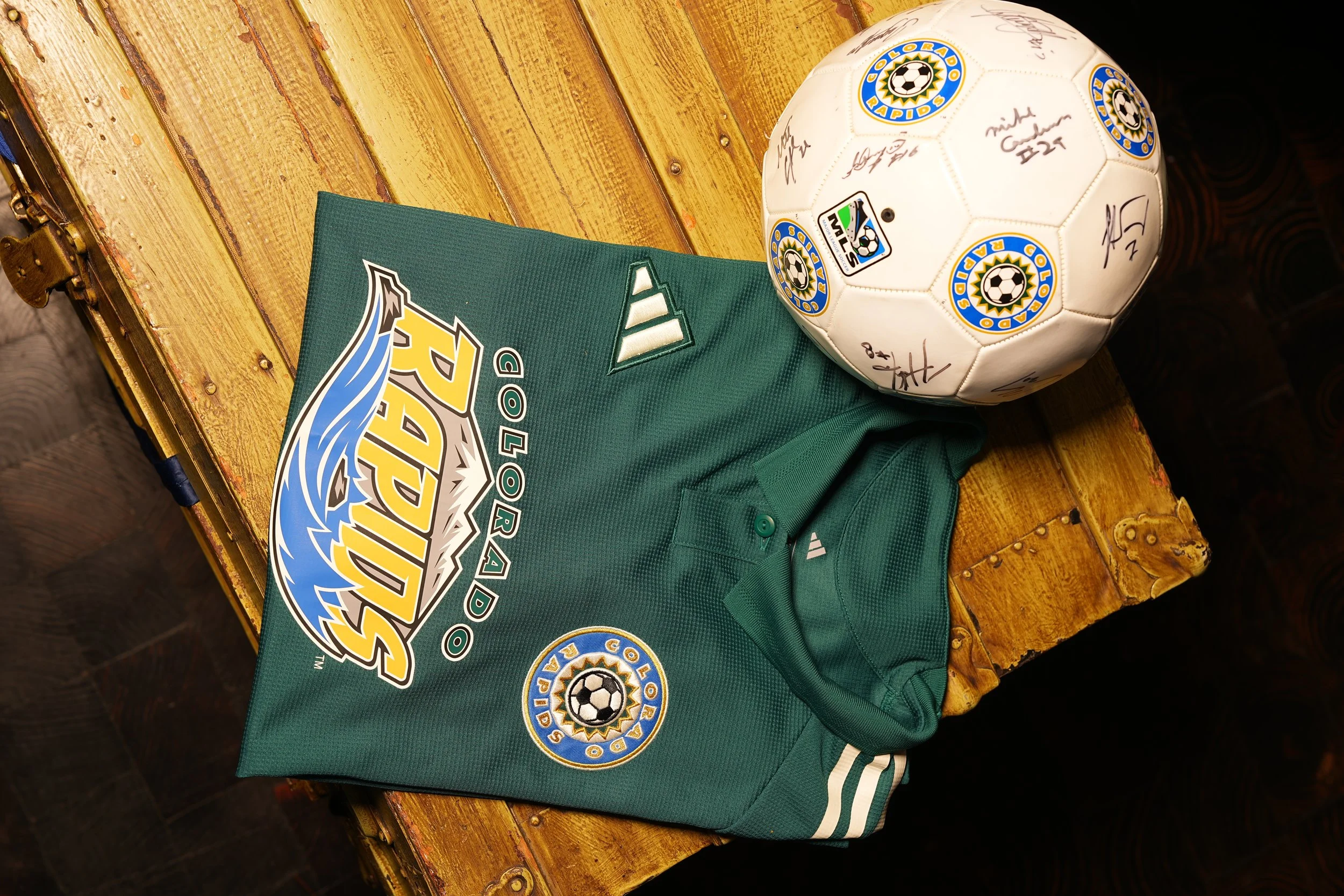 A green Colorado Rapids soccer jersey, a white soccer ball with signatures and logo, both on a wooden surface.