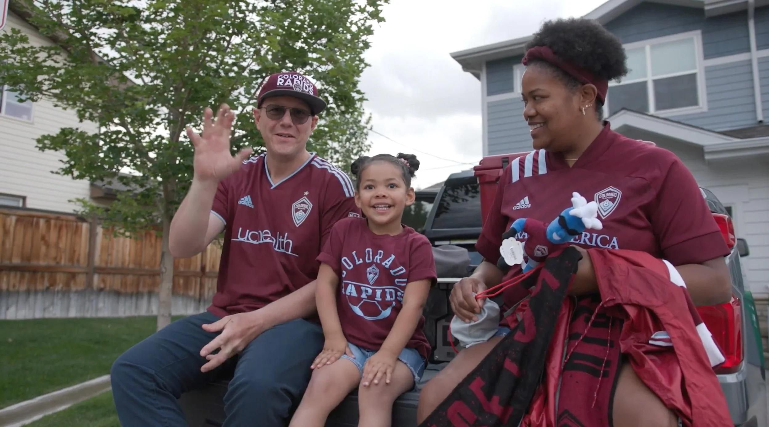 A family of three sitting on the back of a pickup truck outdoors, wearing Colorado Rapids soccer jerseys. The man is waving and wearing sunglasses and a cap, the young girl in the middle is smiling, and the woman on the right is holding soccer plush toys and smiling.