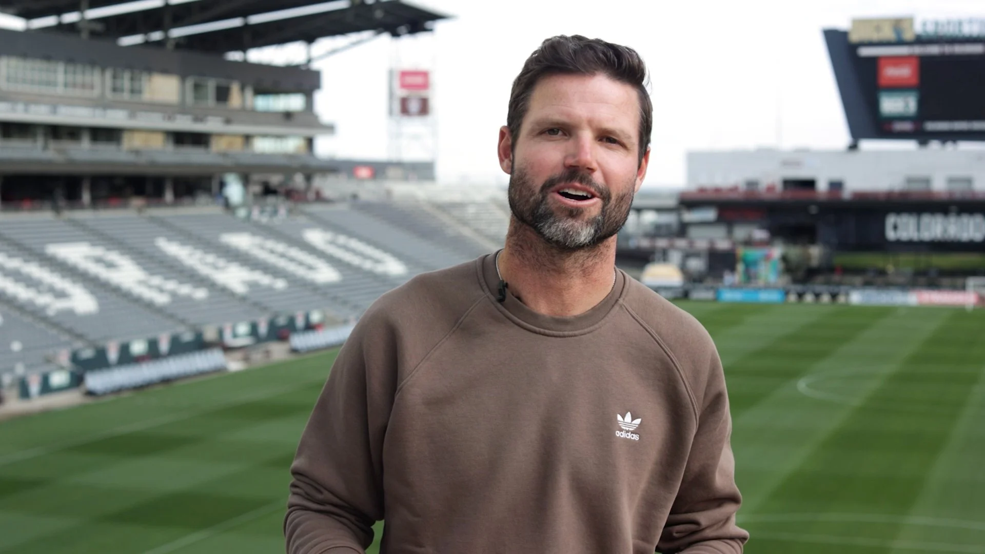 Man with brown hair and beard wearing a brown Adidas sweatshirt standing on a football field in a stadium.
