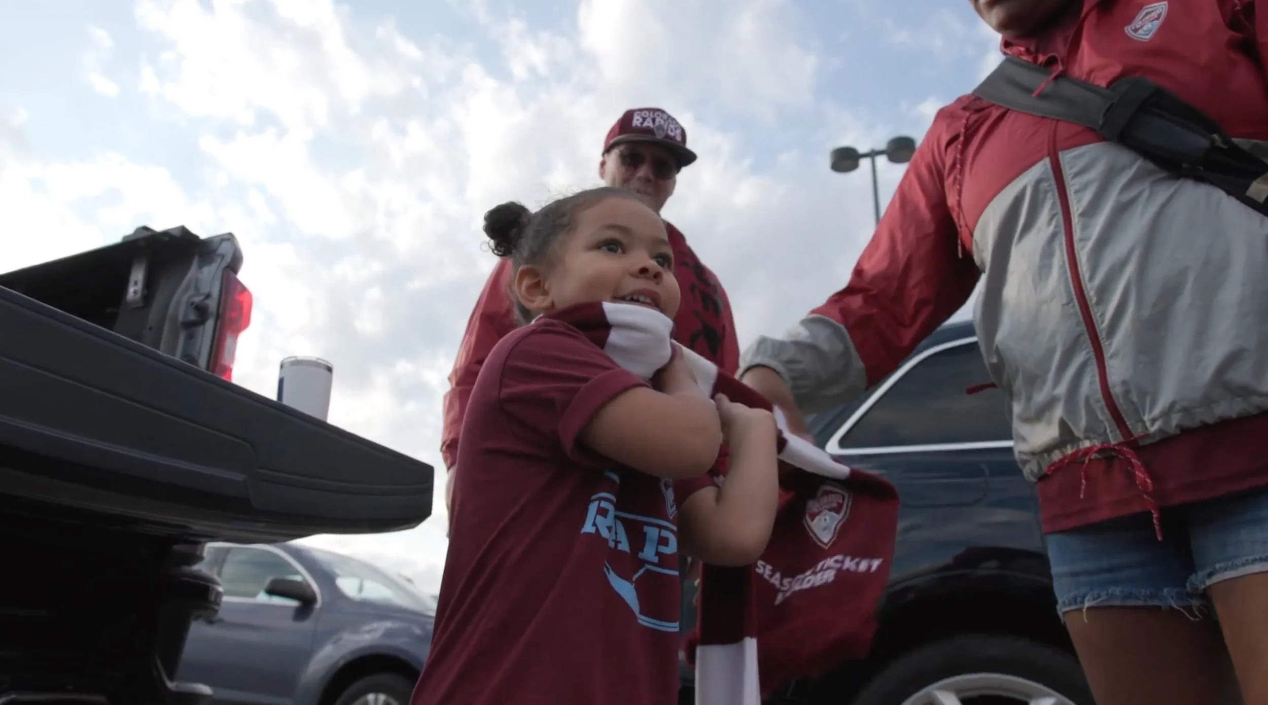 A young girl with braided hair is smiling and holding a scarf, standing next to a woman. They are in a parking lot with cars visible in the background. The girl is wearing a maroon T-shirt with a logo, and the woman is wearing a red jacket and denim shorts. The scene is outdoors during the daytime with partly cloudy skies.