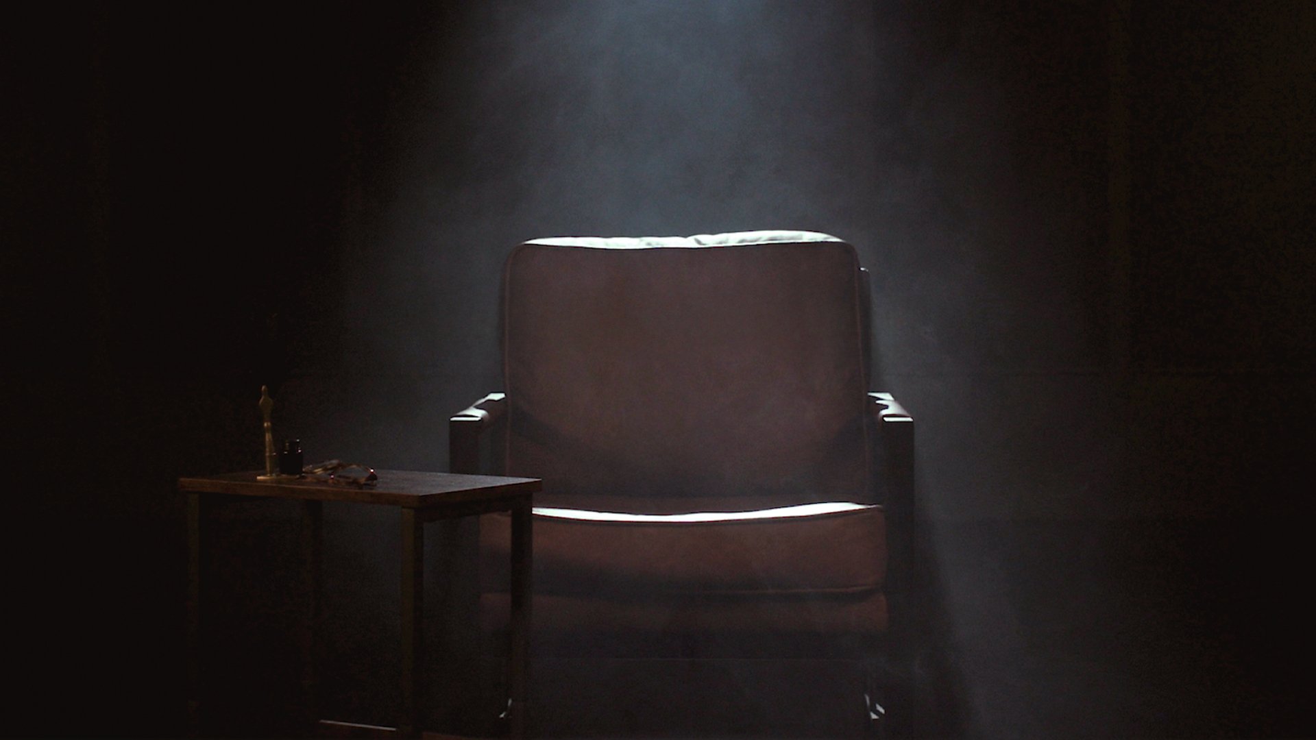 A dark, empty room with a single spotlight illuminating an empty armchair and a small table beside it with a bottle, some objects, and a small statue.