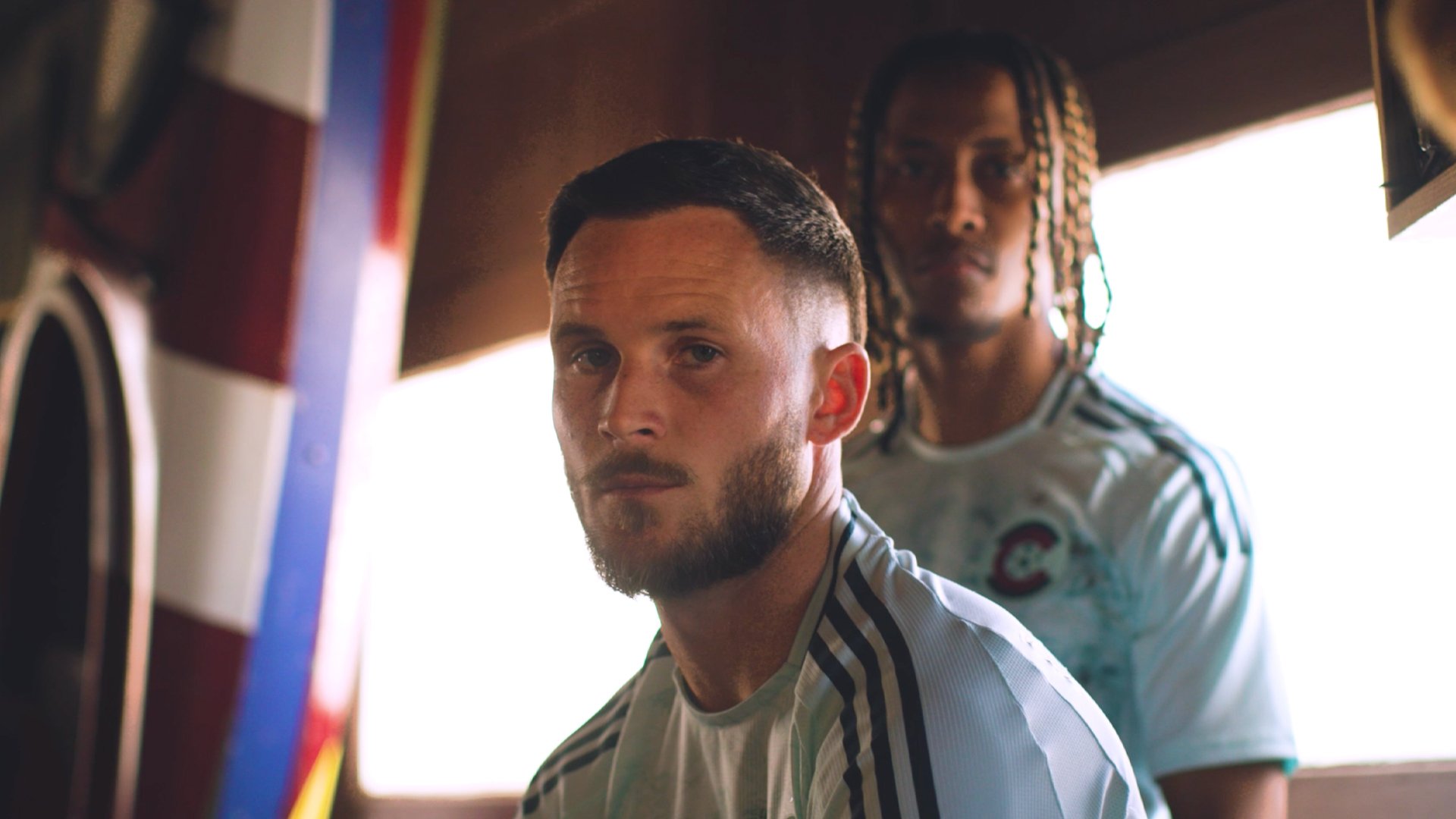 Two men in sports jerseys, one in front with a beard and short hair, looking directly at camera, the other in back with braided hair, looking slightly away, inside a room with sunlight coming through a window.