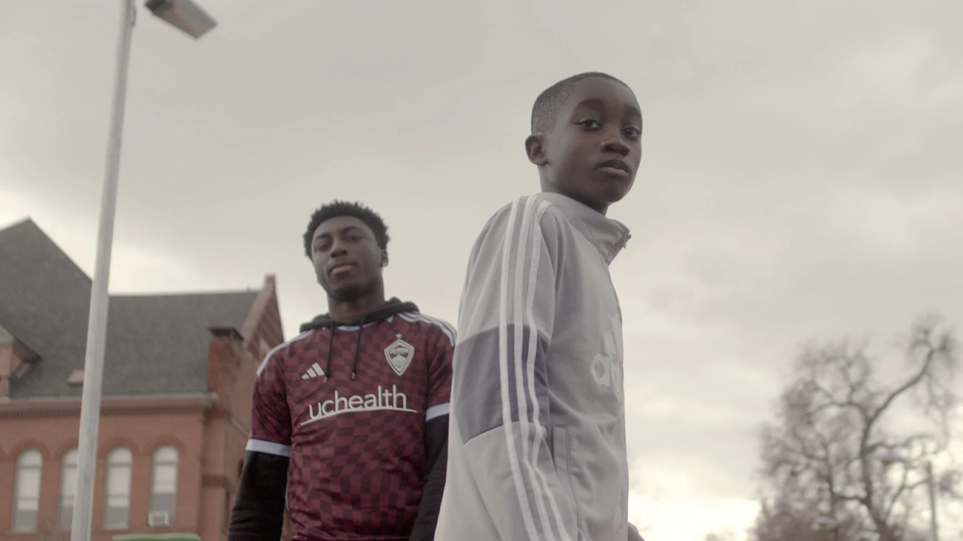 Two young boys standing outdoors on a cloudy day, one wearing a white jacket and the other wearing a maroon and white sports jersey, with a building and leafless trees in the background.