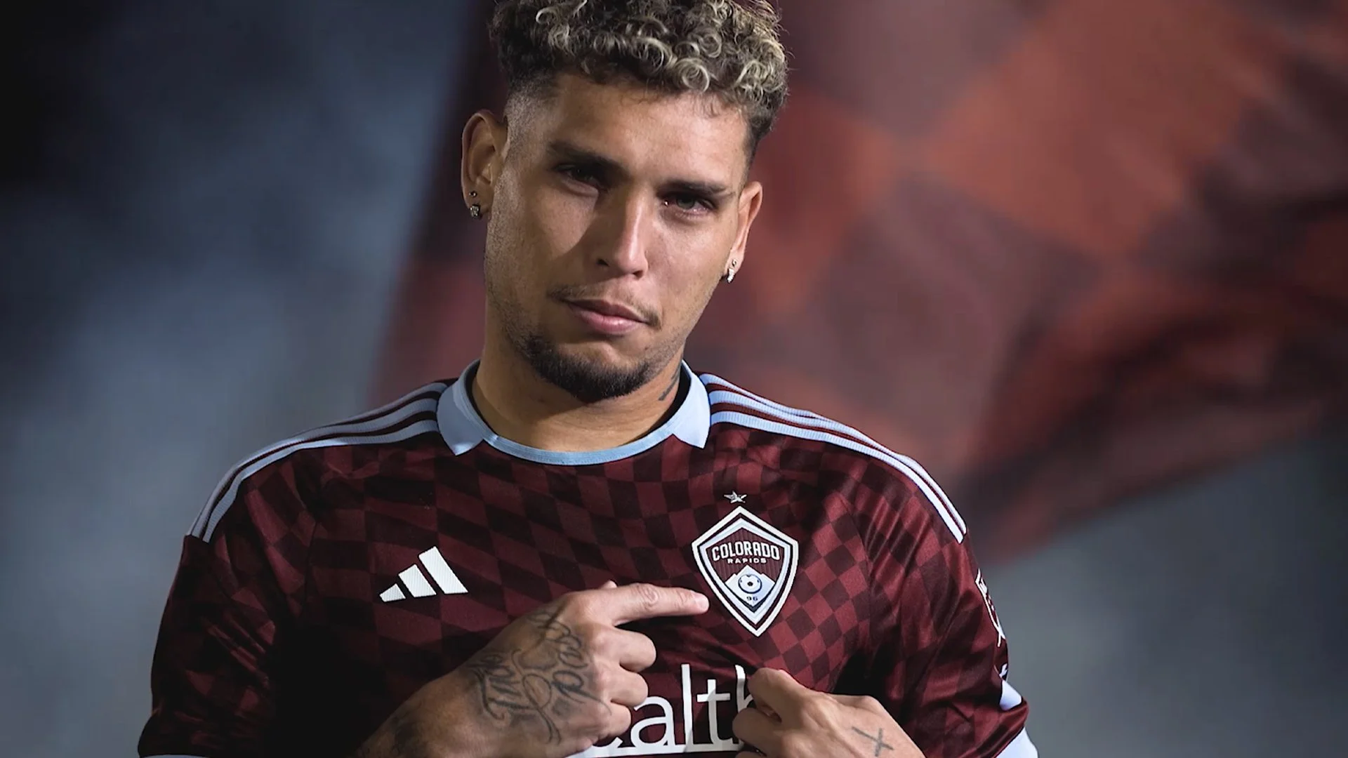 A male soccer player with short curly hair and earrings, wearing a maroon and black Colorado Rapids soccer jersey, points to the team logo on his chest.