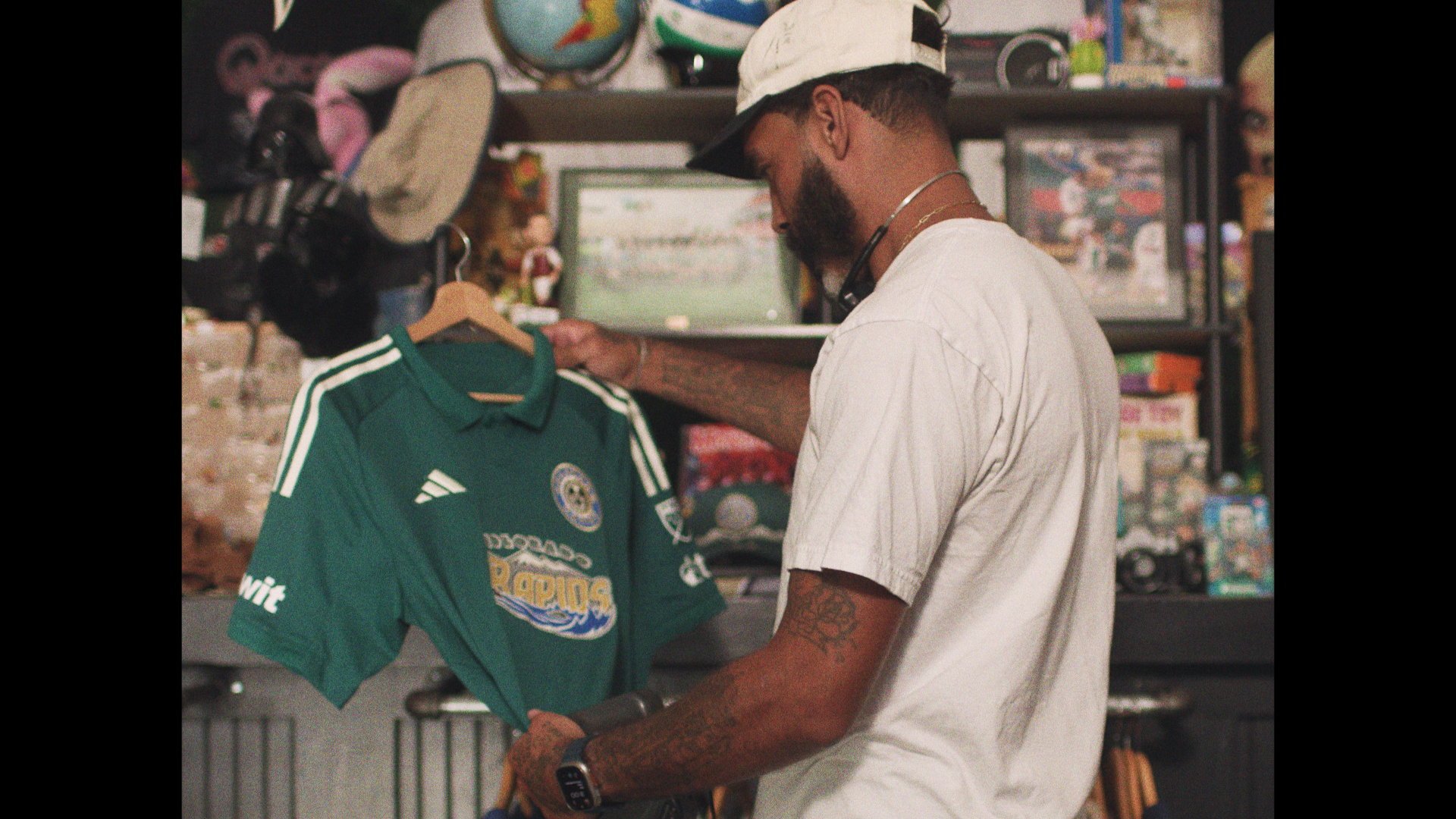 A man with a beard and tattoos is looking at a green sports jersey with white stripes, hanging on a hanger, in a store filled with various collectibles and memorabilia.