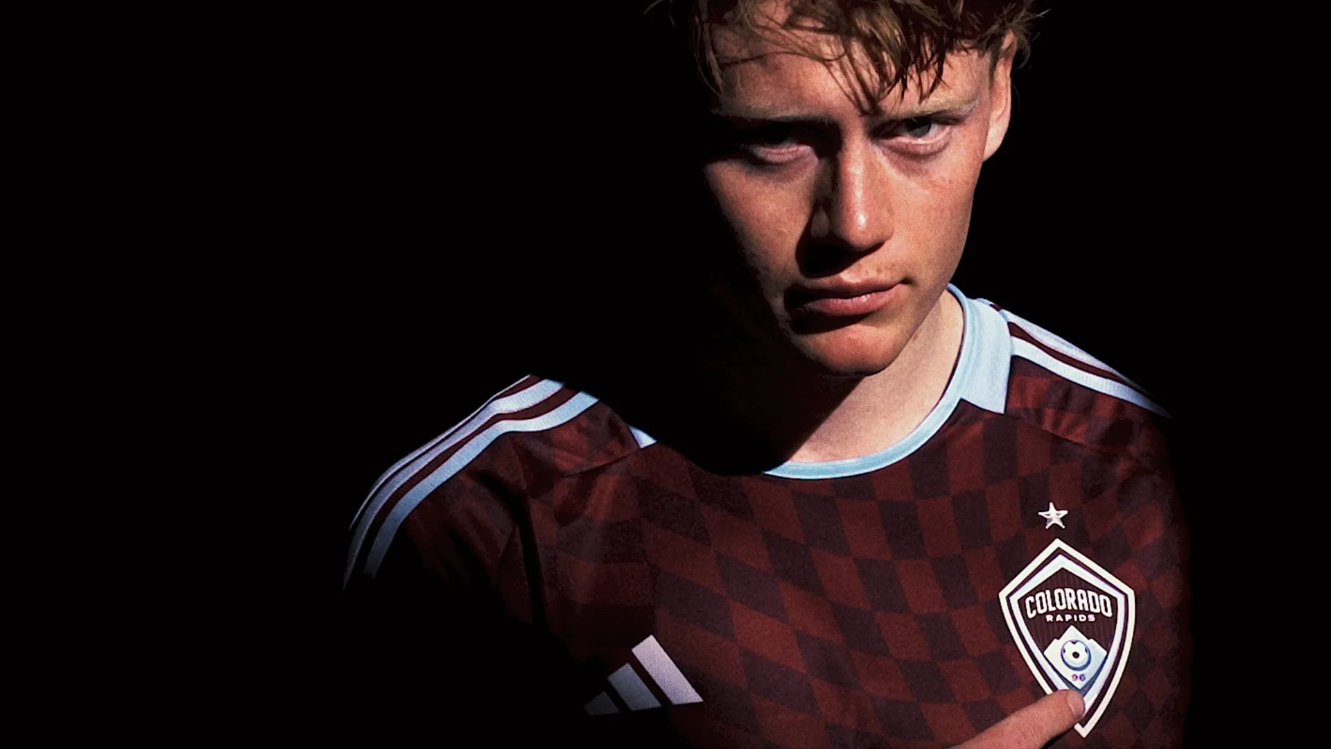 A young man with light skin and blond hair looking directly at the camera, wearing a maroon soccer jersey with white accents and the Colorado Rapids logo.
