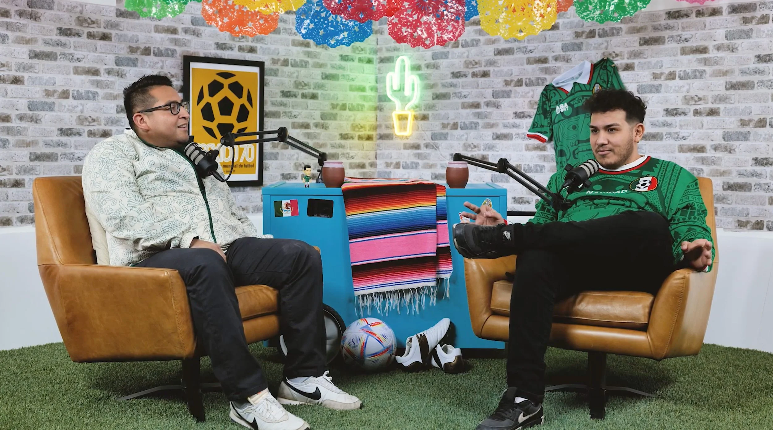 Two men are sitting on tan leather chairs facing each other in a colorful, decorated room with a brick wall background. They have microphones in front of them, suggesting a podcast or interview setting. The room includes soccer memorabilia, a neon cactus light, and festive paper decorations hanging from the ceiling.