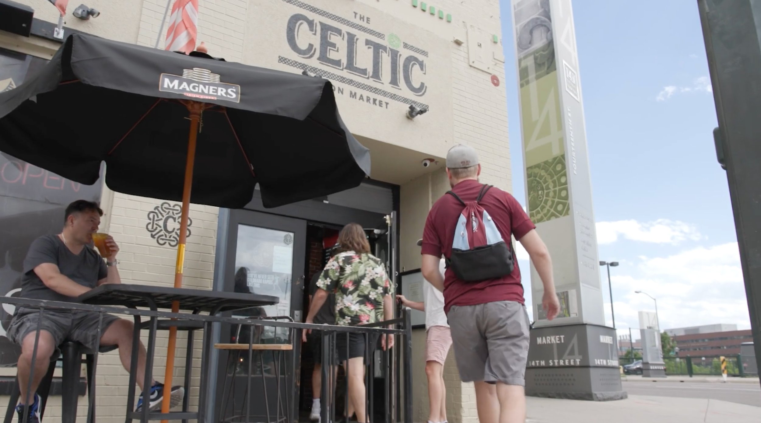 People entering and sitting outside the Celtic Market, which has a sign above the entrance with the name. One person is sitting at a table under a large black umbrella labeled 'Magners', drinking a beverage. Three other people are walking toward the entrance.
