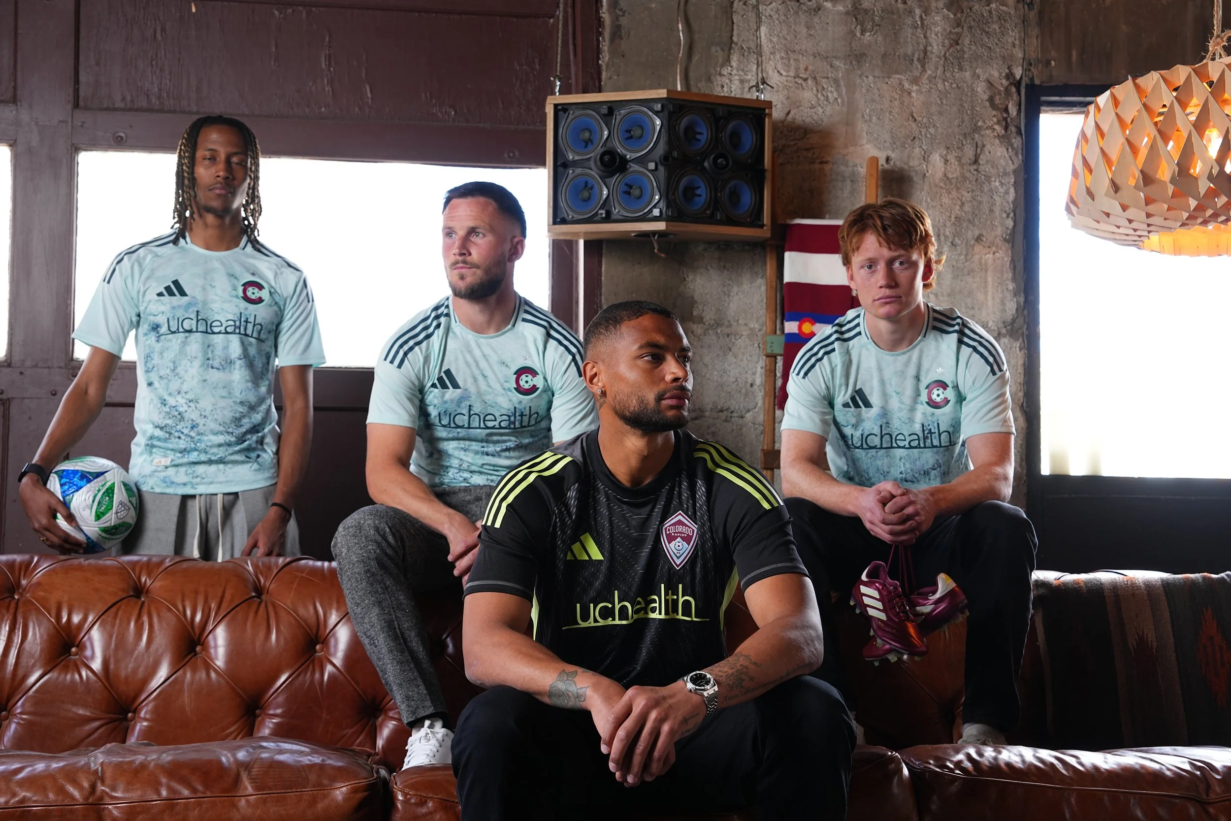 Four soccer players in team jerseys sitting and standing on a leather couch in a rustic room with large windows, speakers, and Colorado flag. One player holds a soccer ball, others have soccer shoes visible.