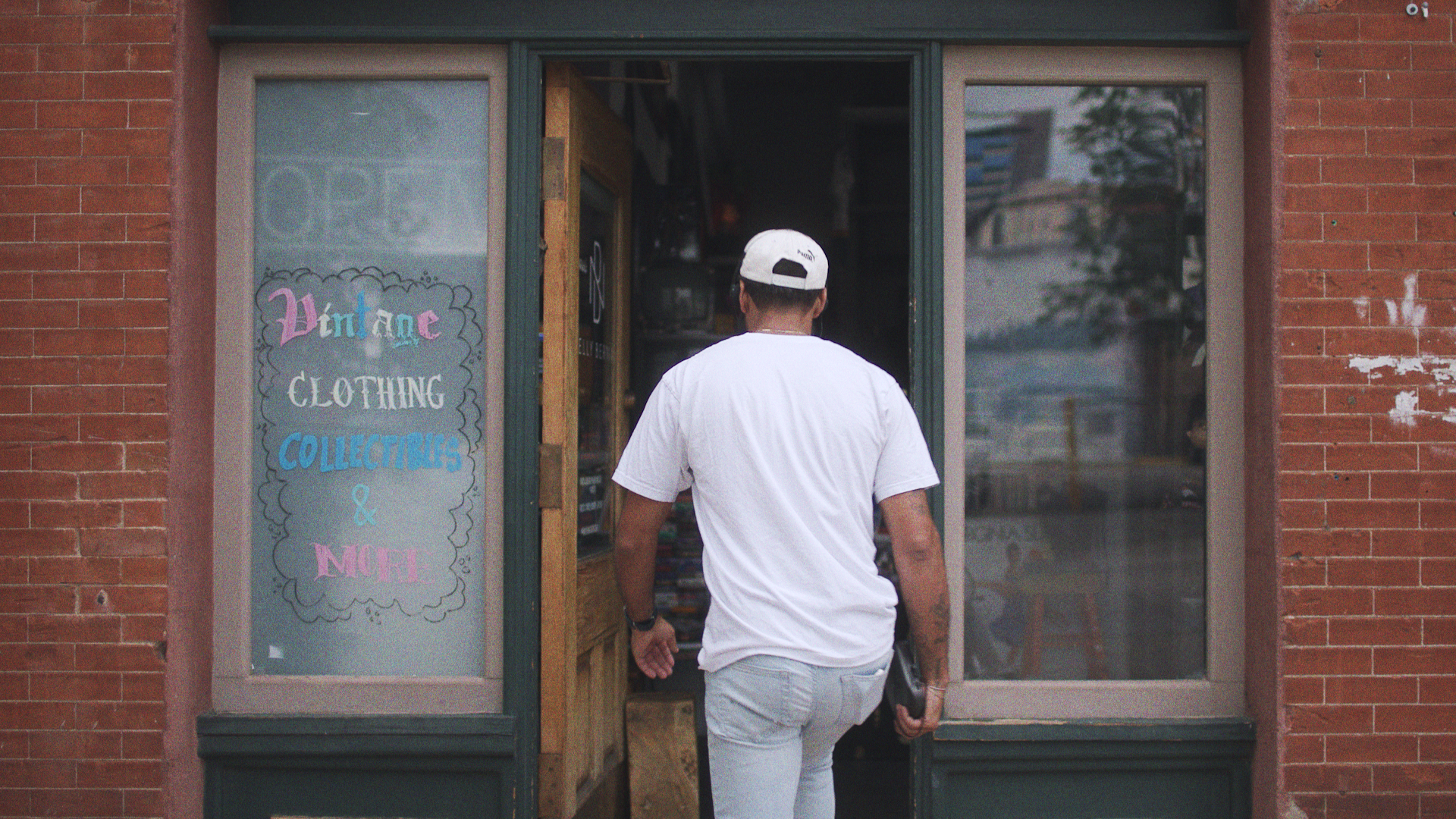 A man wearing a white t-shirt and a white cap walks into a store with a sign that reads 'Vintage Clothing, Collectibles & More' on the window, next to a brick building.