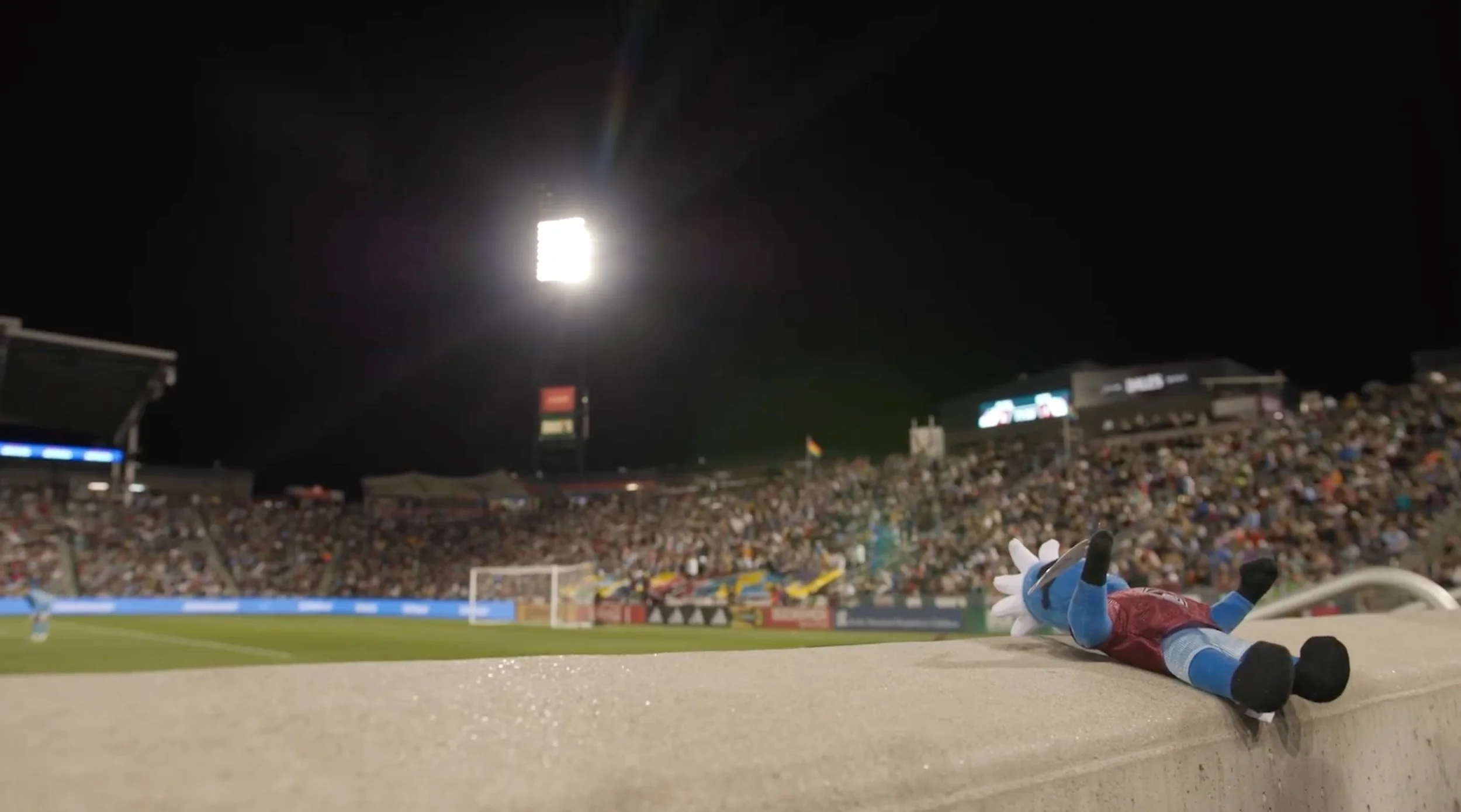 A stuffed Sonic the Hedgehog toy lies on a ledge overlooking a soccer stadium filled with spectators at night. Bright stadium lights illuminate the field.
