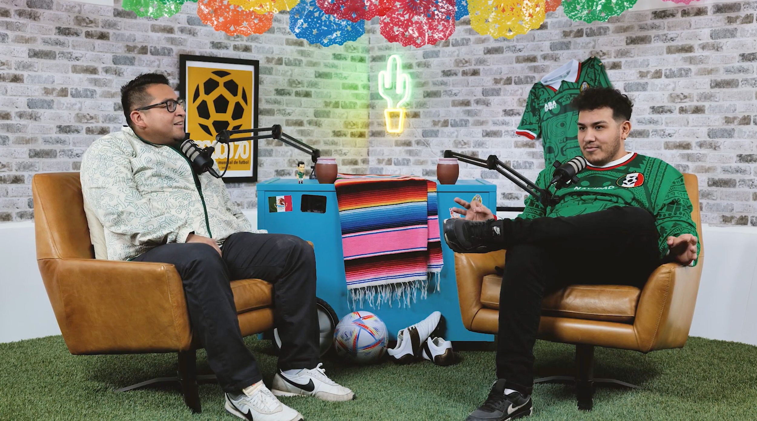 Two men sitting in chairs in a colorful, casual studio setting with a brick wall background, microphones, and soccer-themed decor, engaging in conversation.