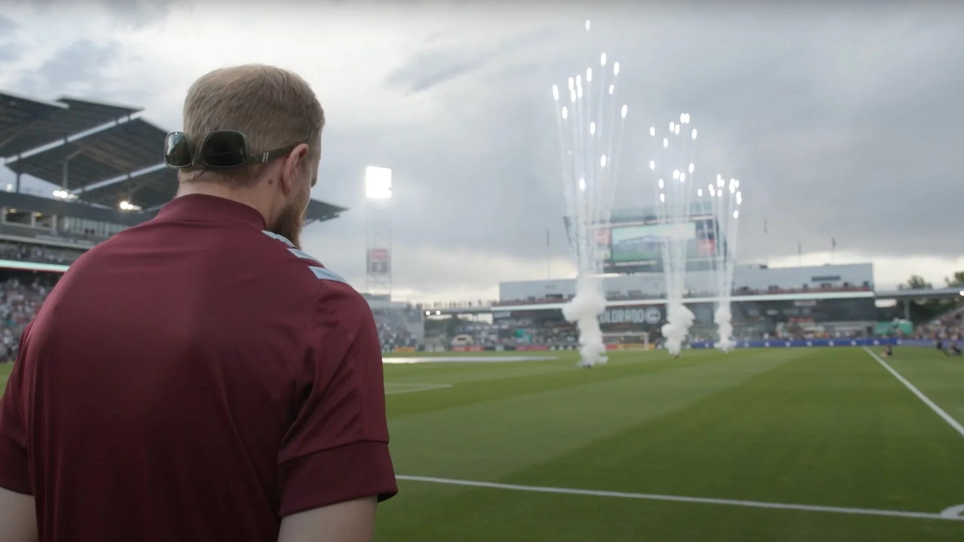 A man in a maroon sports uniform with headphones around his neck watching fireworks on a sports field.