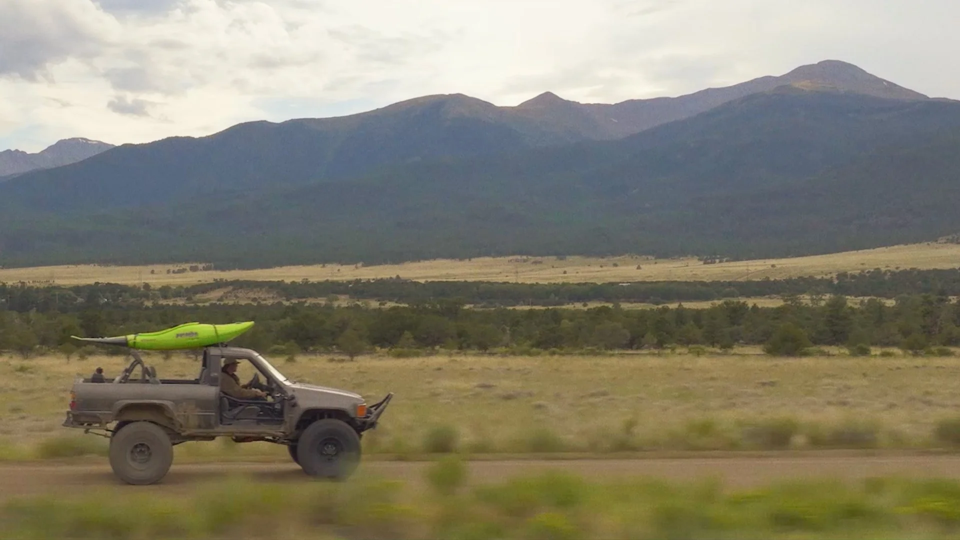 A rugged pickup truck driving through a desert landscape with mountains in the background. The truck has a kayak mounted on its roof, and there is a person sitting in the driver's seat.