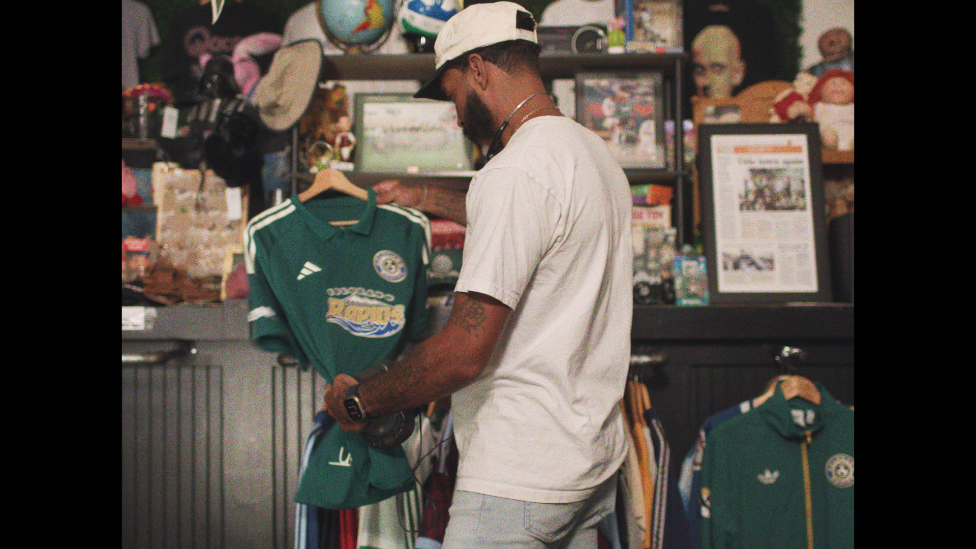 A man with tattoos wearing a white striped shirt and a backwards cap shopping for a green soccer jersey in a store filled with sports merchandise, including hats, framed pictures, and dolls.