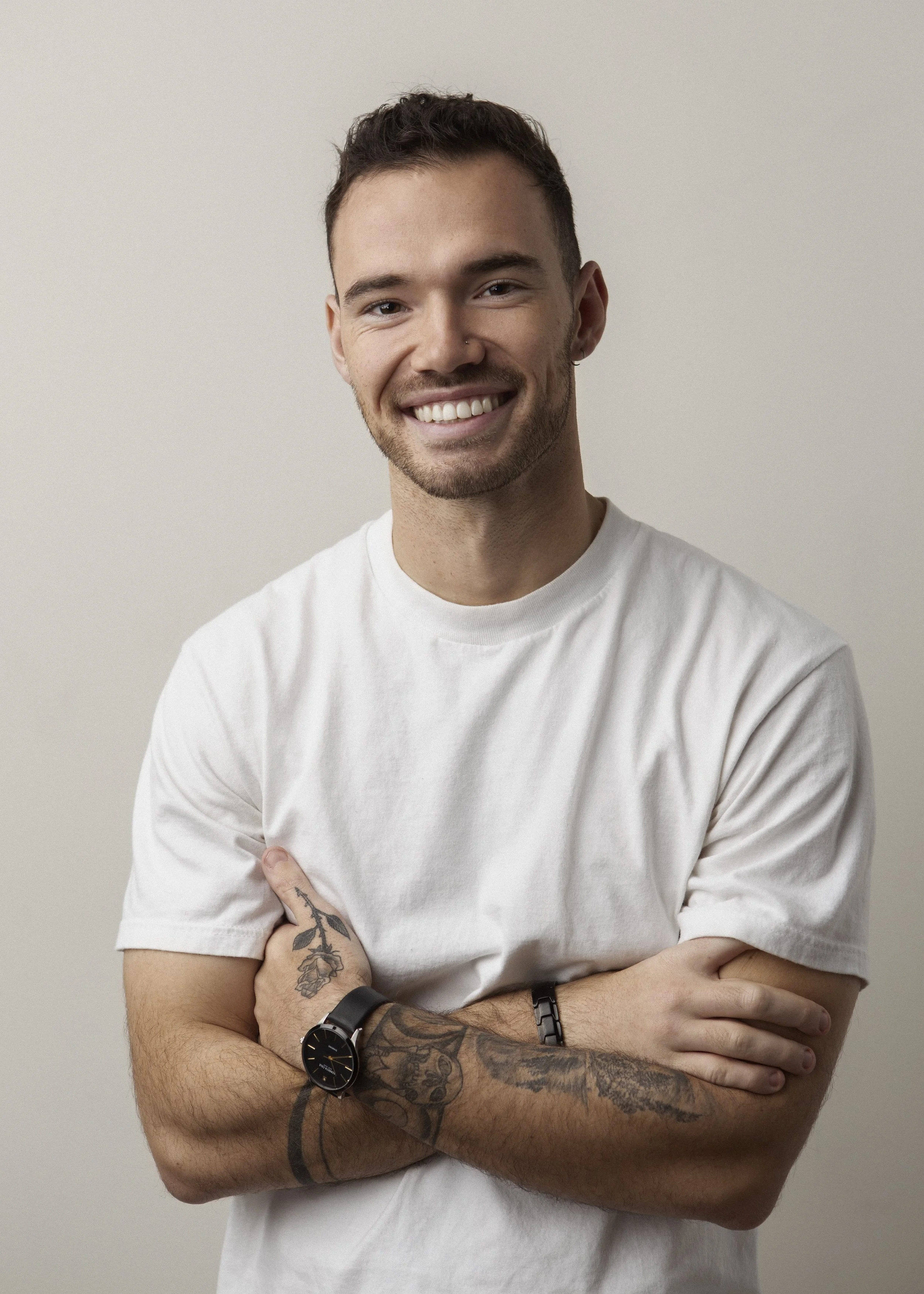 A young man with short dark hair, a beard, and tattoos on his left arm, smiling and crossing his arms, wearing a white t-shirt and a black watch, against a plain background.