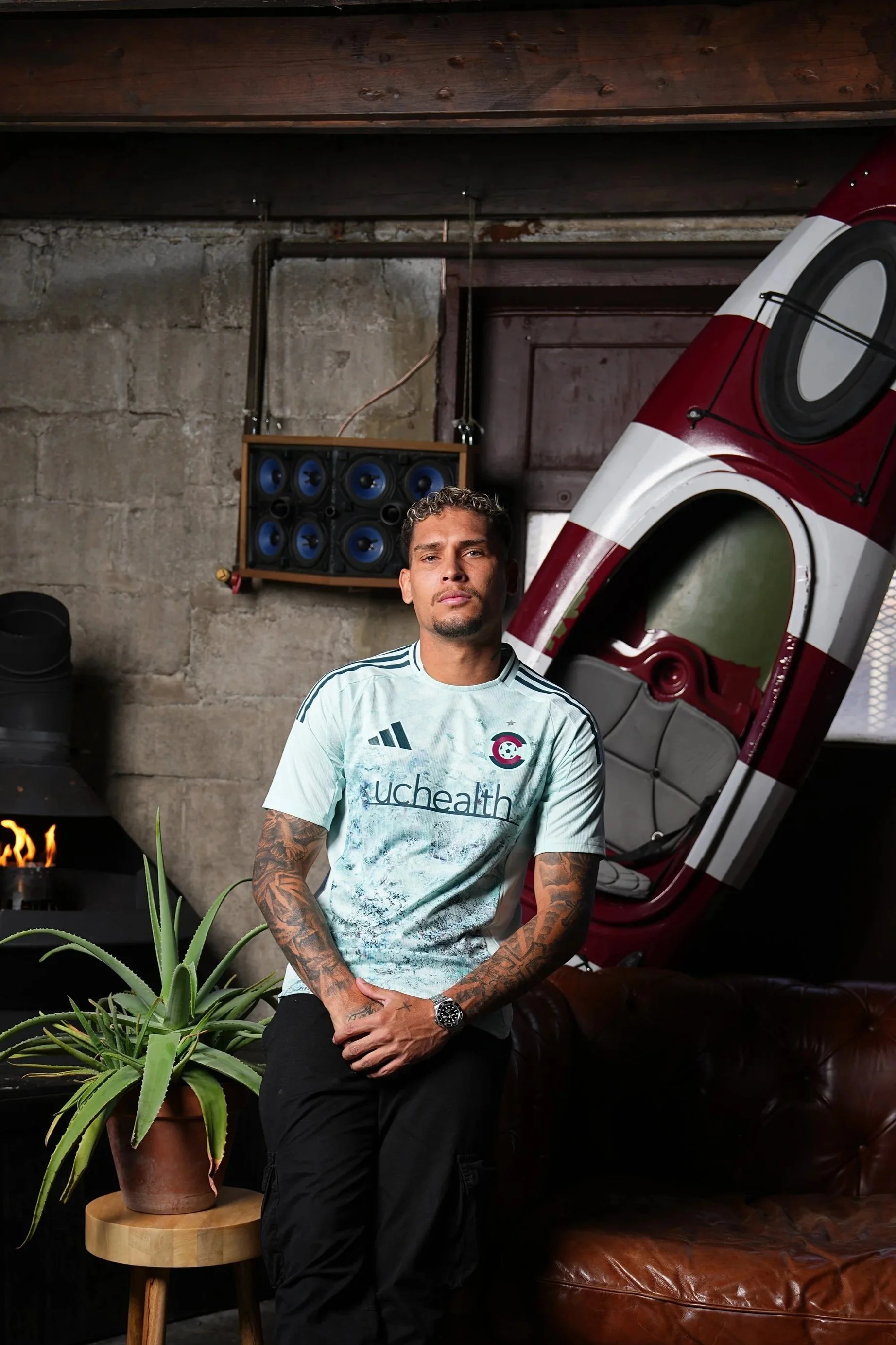 A man with tattoos on his arms standing indoors against a concrete wall, with a maroon and white kayak behind him, a potted aloe vera plant on a stool to his left, and a wall-mounted speaker system in the background.