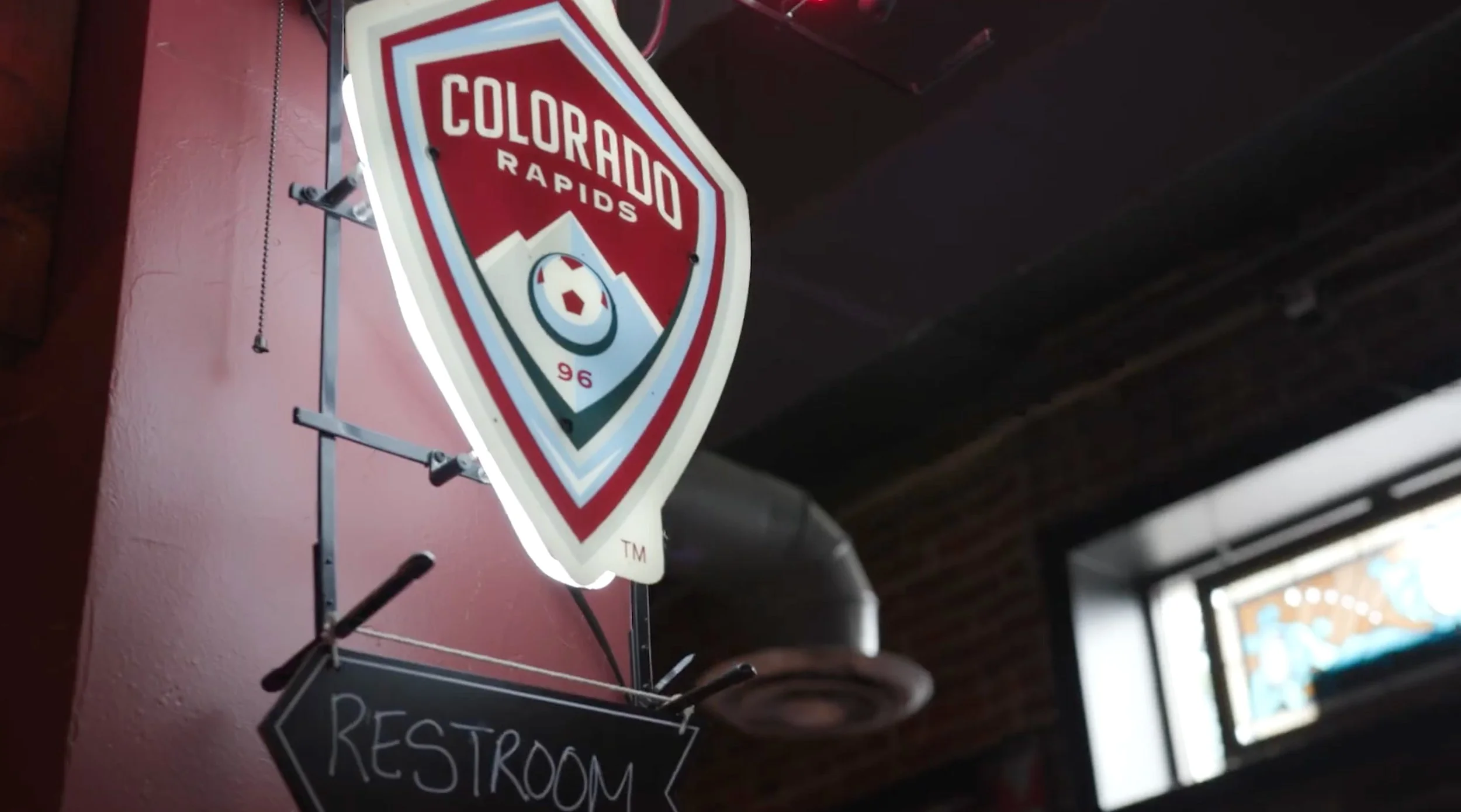 A neon sign of the Colorado Rapids soccer team logo hanging on a pink wall, with a black sign underneath reading 'RESTROOM' in chalk.