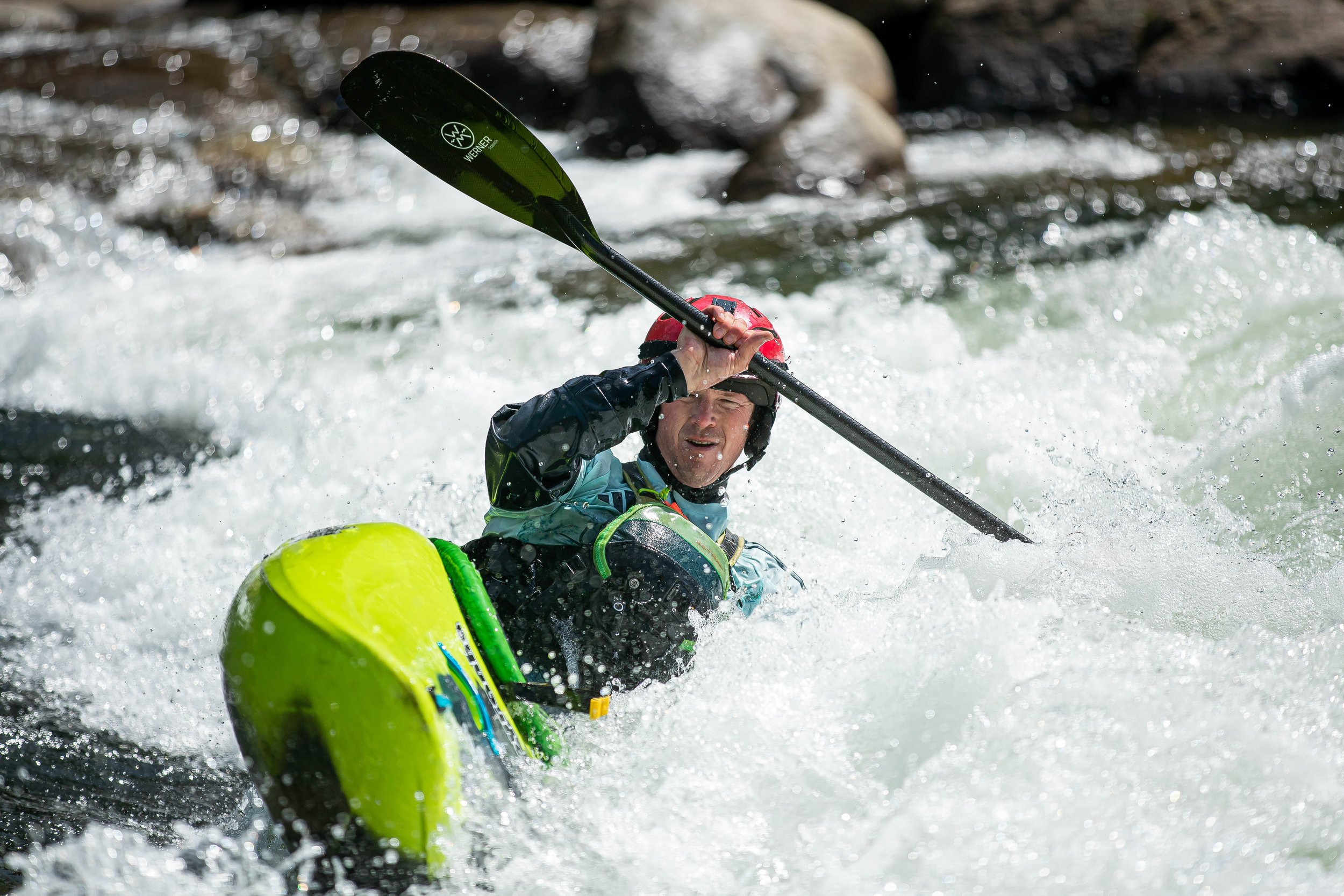 A man kayaking in a rapid river, wearing a red helmet and black waterproof gear, holding a black paddle, with churning water around him.
