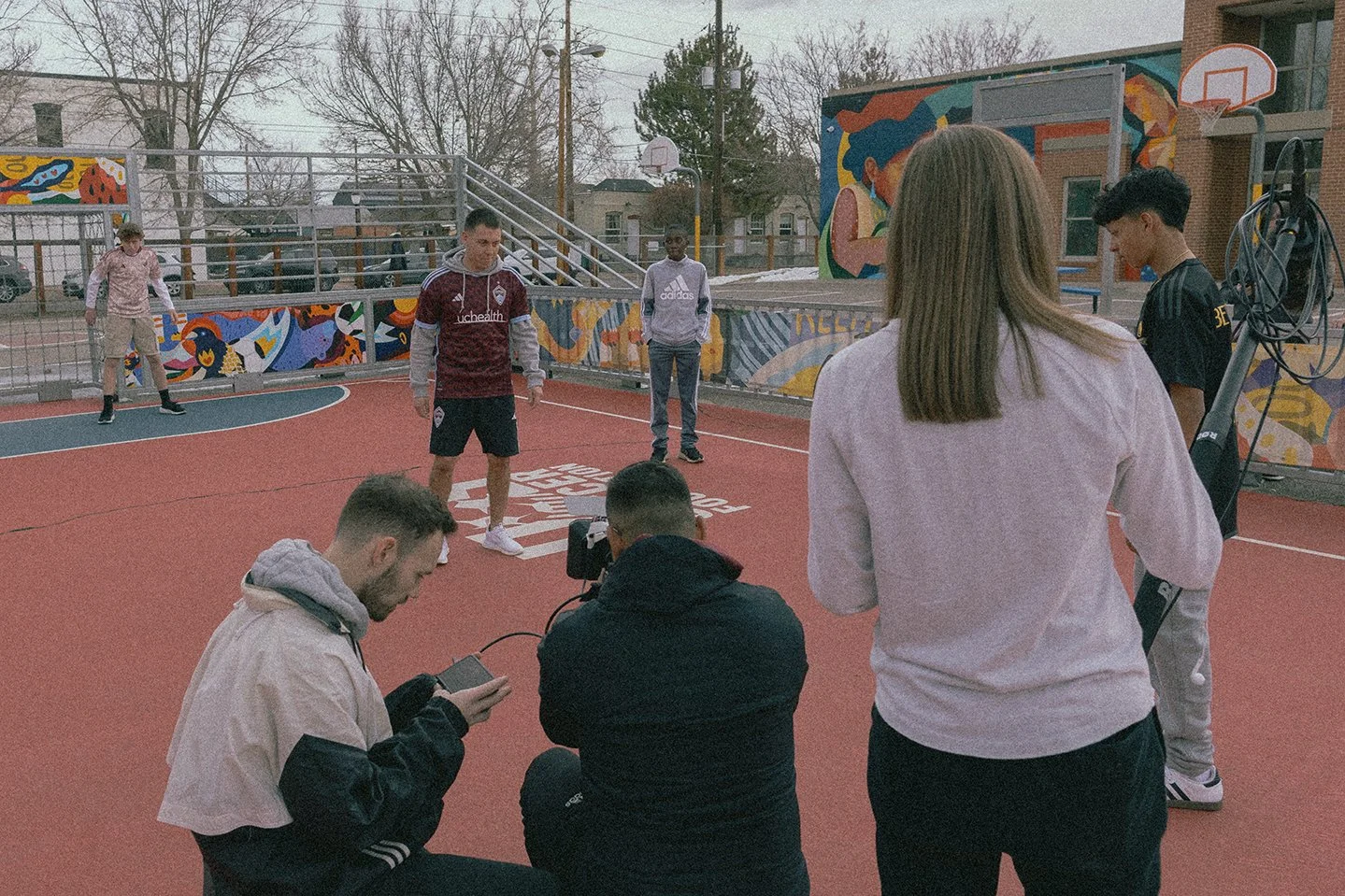 Group of six young people on an outdoor basketball court, some sitting and some standing, during a filming or photoshoot session.