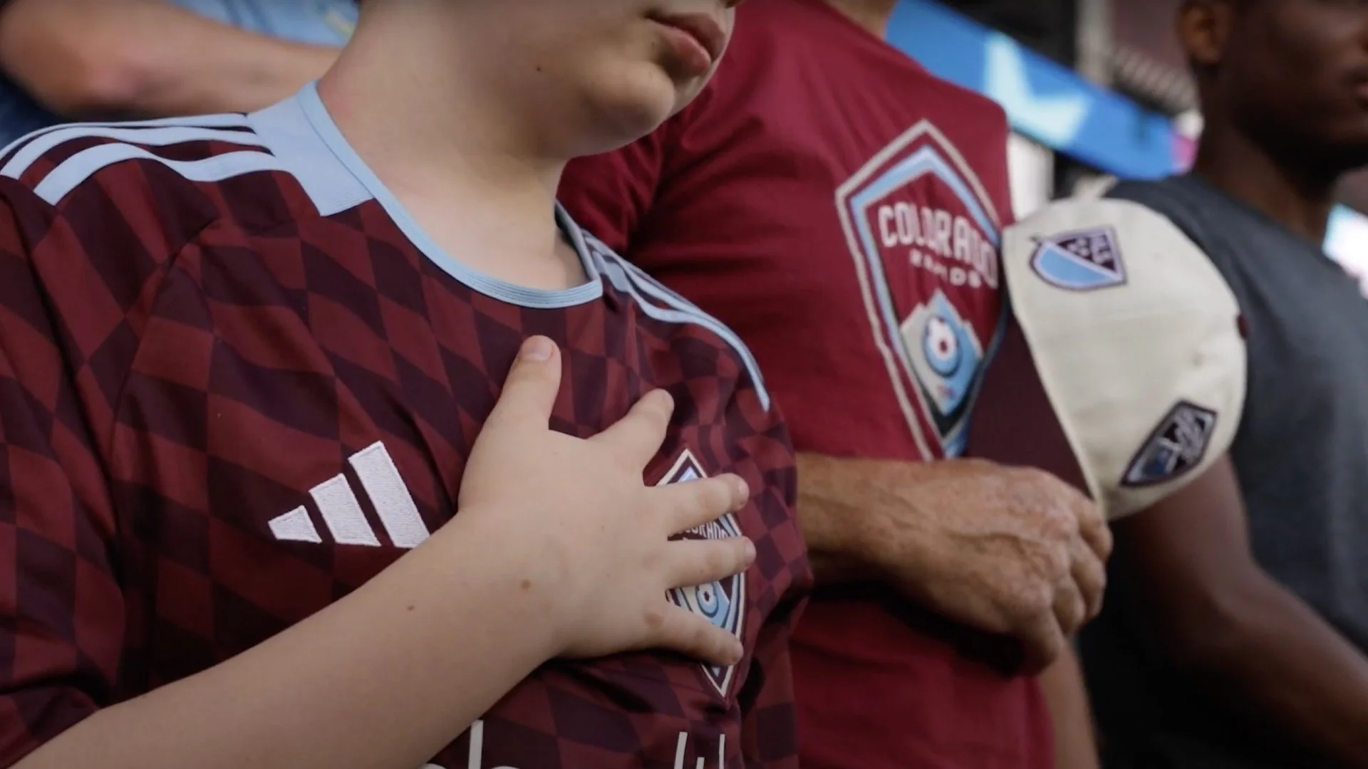A young person with their right hand over their chest, wearing a maroon jersey with white and light blue accents, standing among others during a moment of silence or prayer.