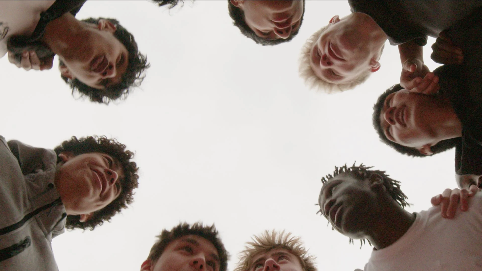 A diverse group of people standing in a circle outdoors, looking down at the camera positioned in the center, with their heads encircling the sky.