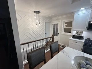 Interior view of a modern kitchen and dining area with white cabinets, a marble countertop, a window, and a staircase with wooden railing.