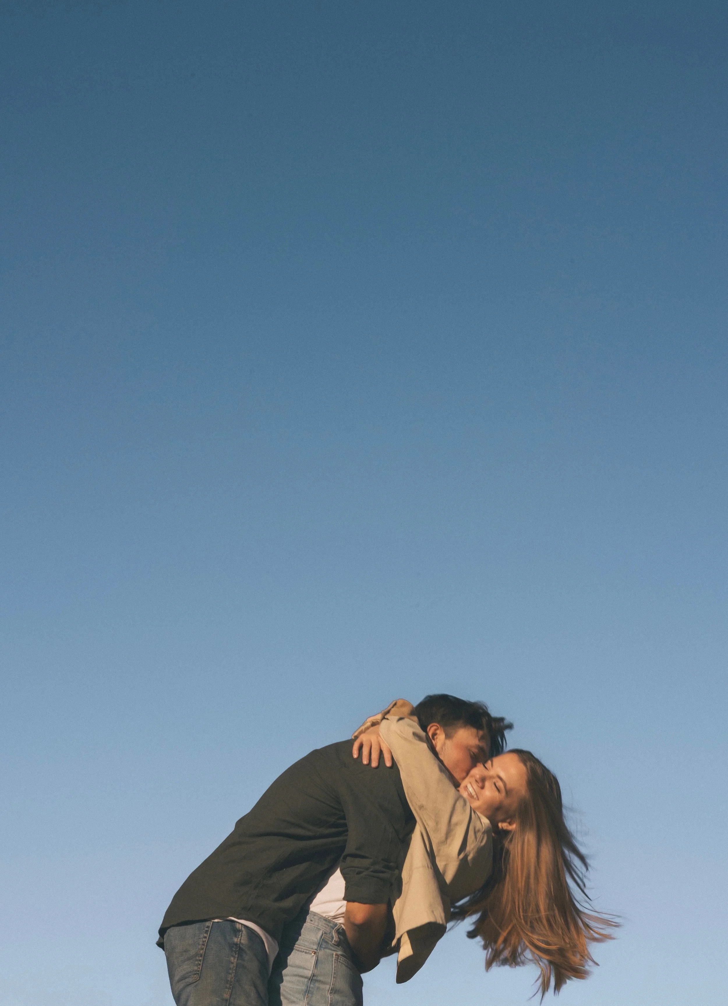 A young couple is outdoors, embracing and sharing a joyful moment against a clear blue sky.