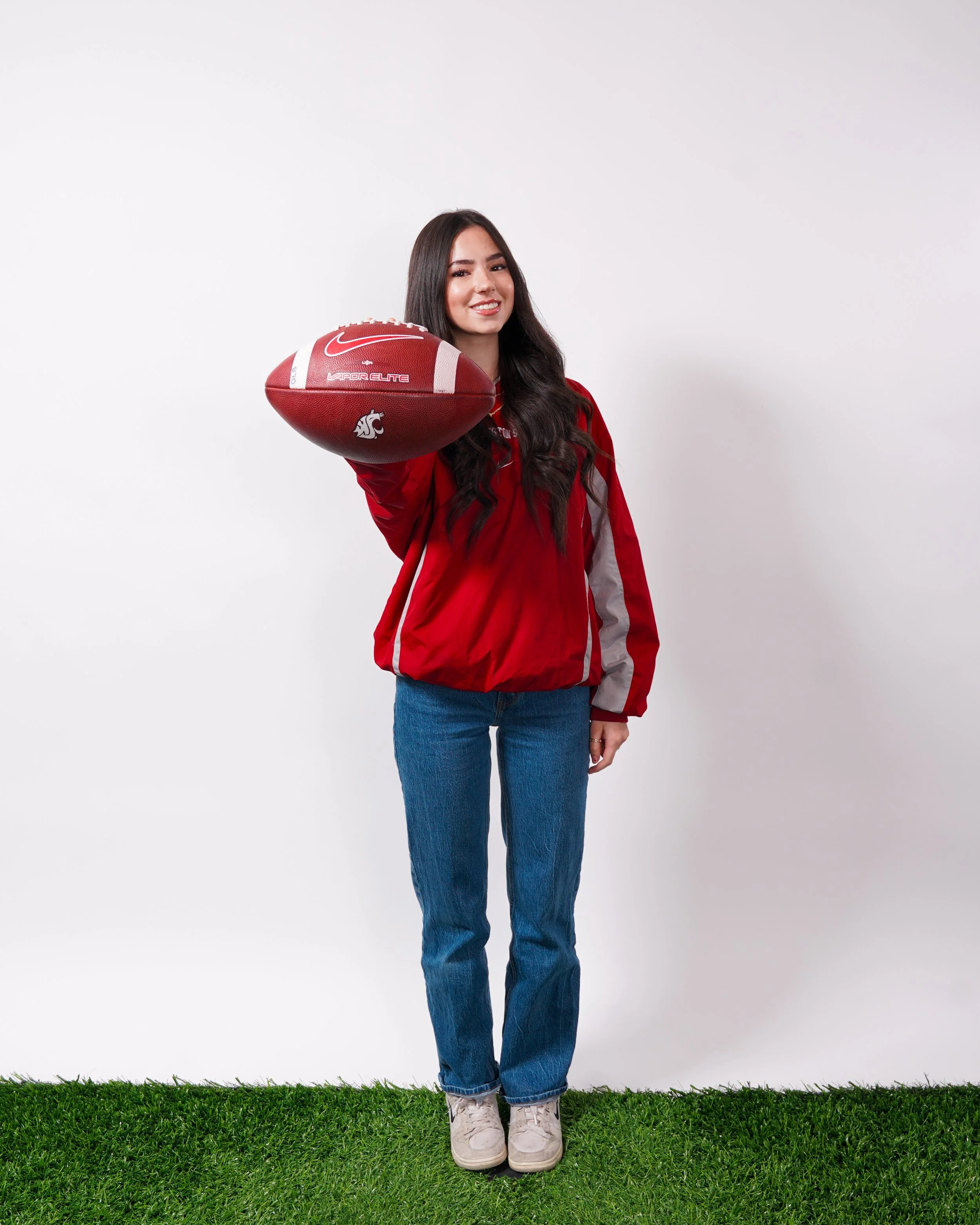 A young woman with long dark hair wearing a red and gray jacket, blue jeans, and white sneakers, standing on green grass against a plain white background, holding a red football.