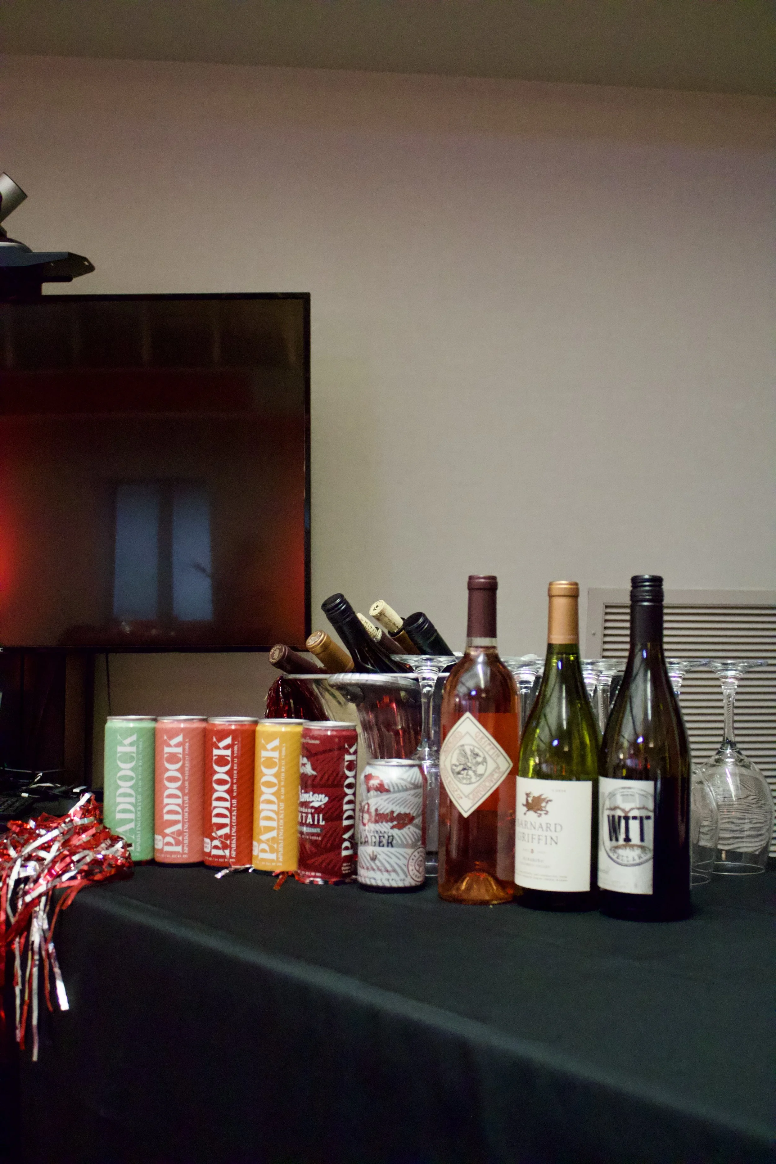 Table with assorted alcoholic beverages, including three cans of flavored bubbly water, a can of sparkling water, several bottles of wine, and empty glasses in the background.