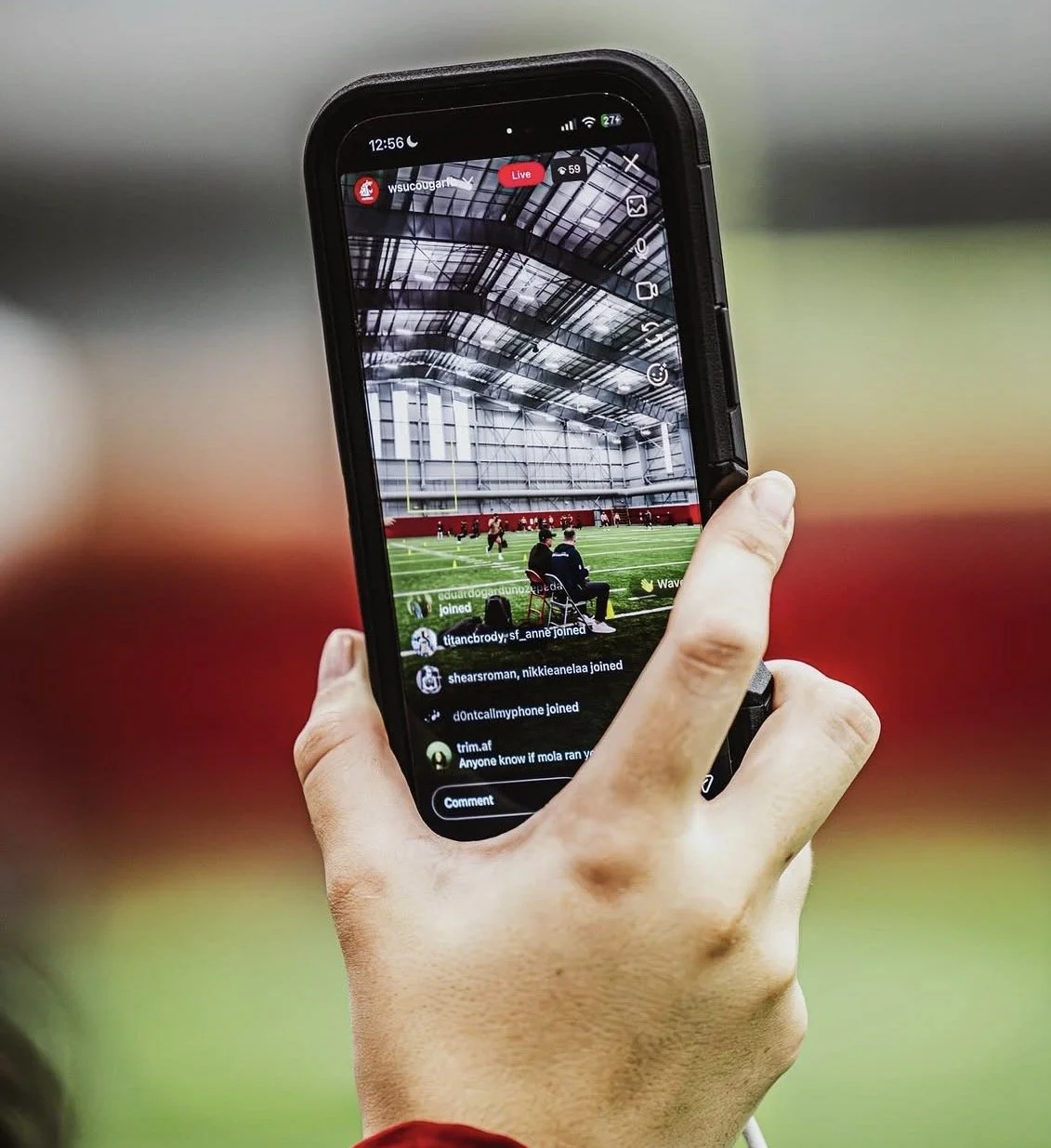 Hand holding a smartphone recording a virtual sports practice inside a large indoor gym with artificial turf and players on the field.