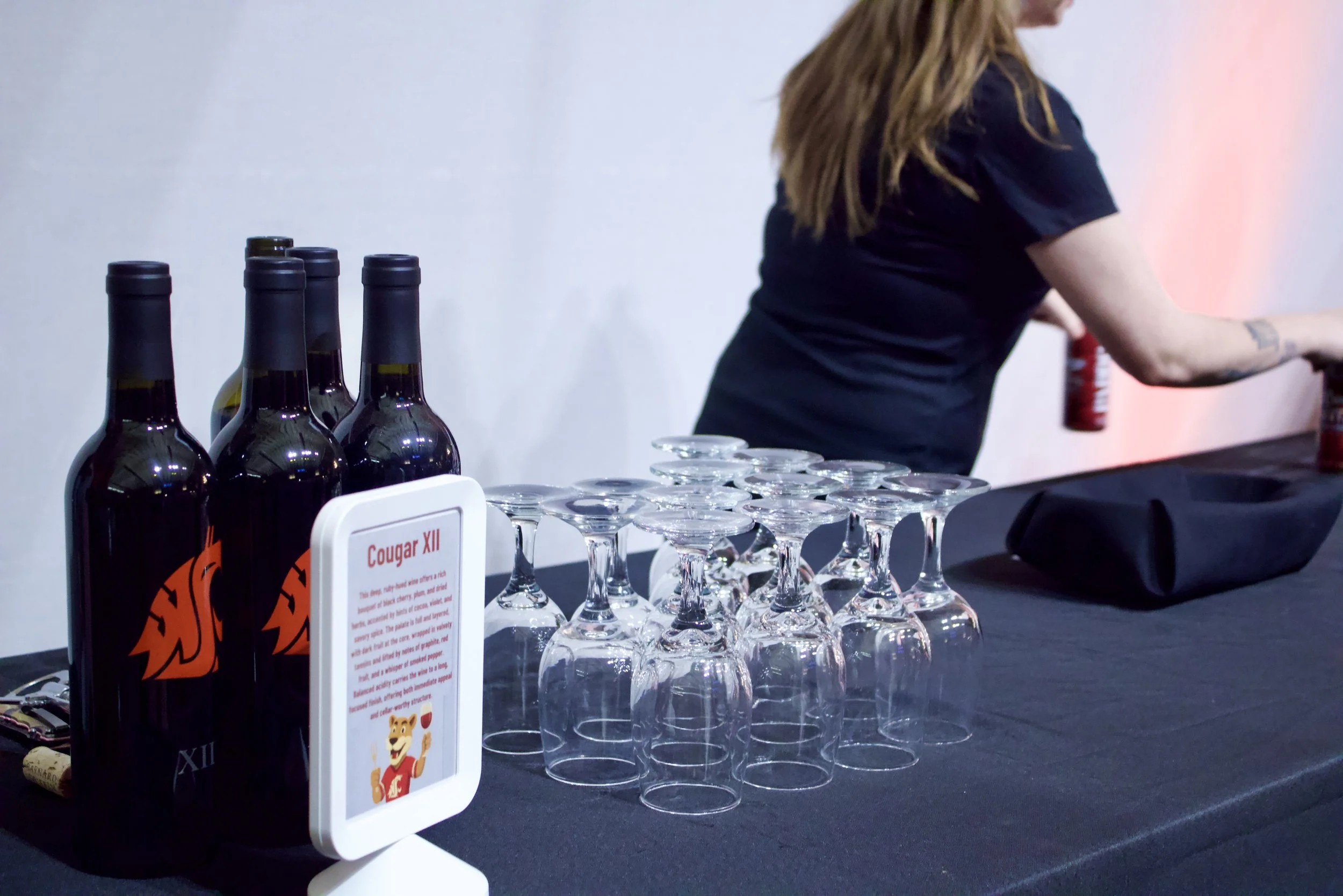 Table with four bottles of Cougar XII wine, upside-down wine glasses, and a sign about Cougar XII, with a woman in black preparing drinks in the background.