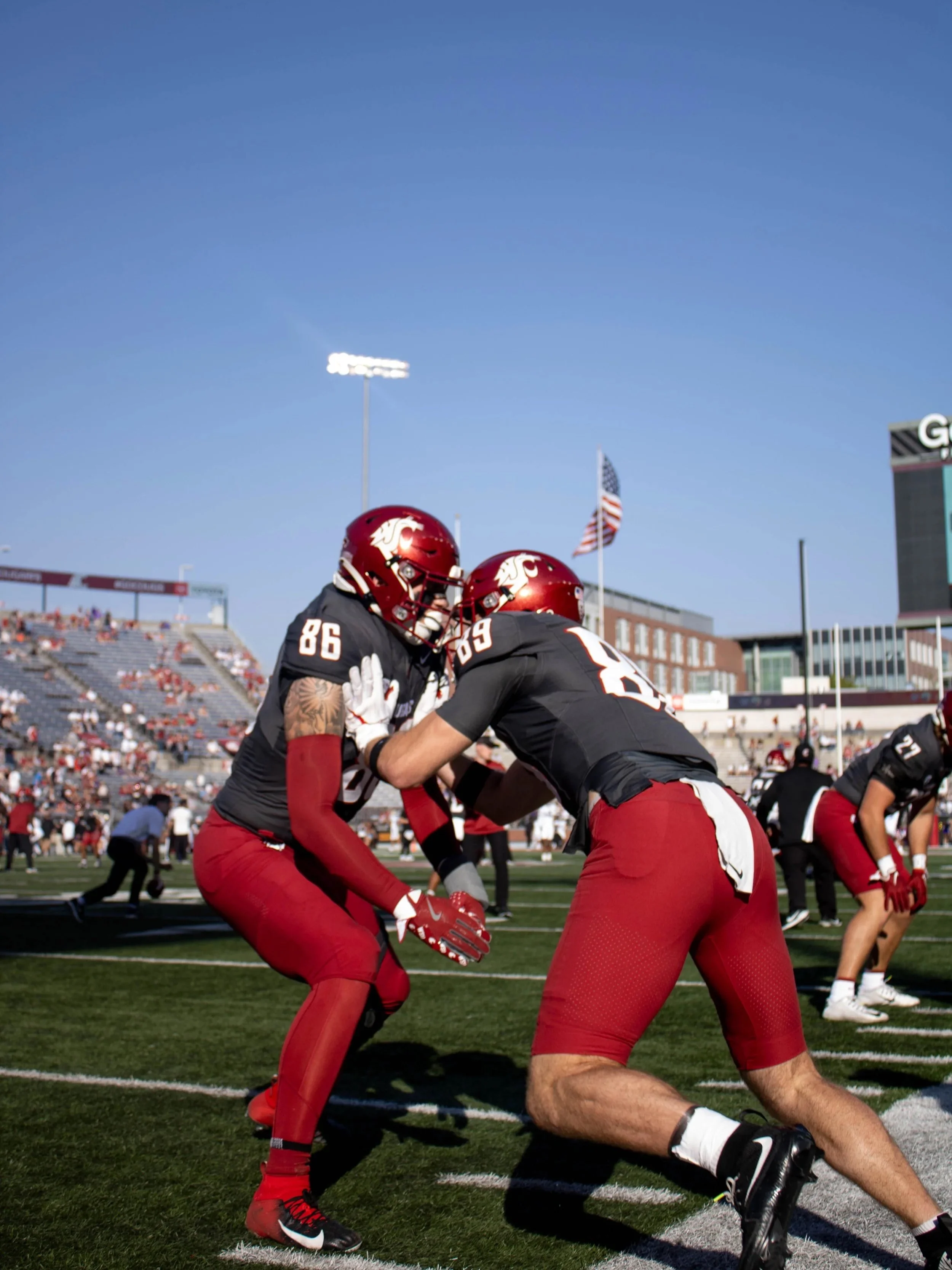 Two football players wearing red and black uniforms on the field, with helmets and pads, engaged in a game during daytime. The background shows a stadium with spectators and flags.