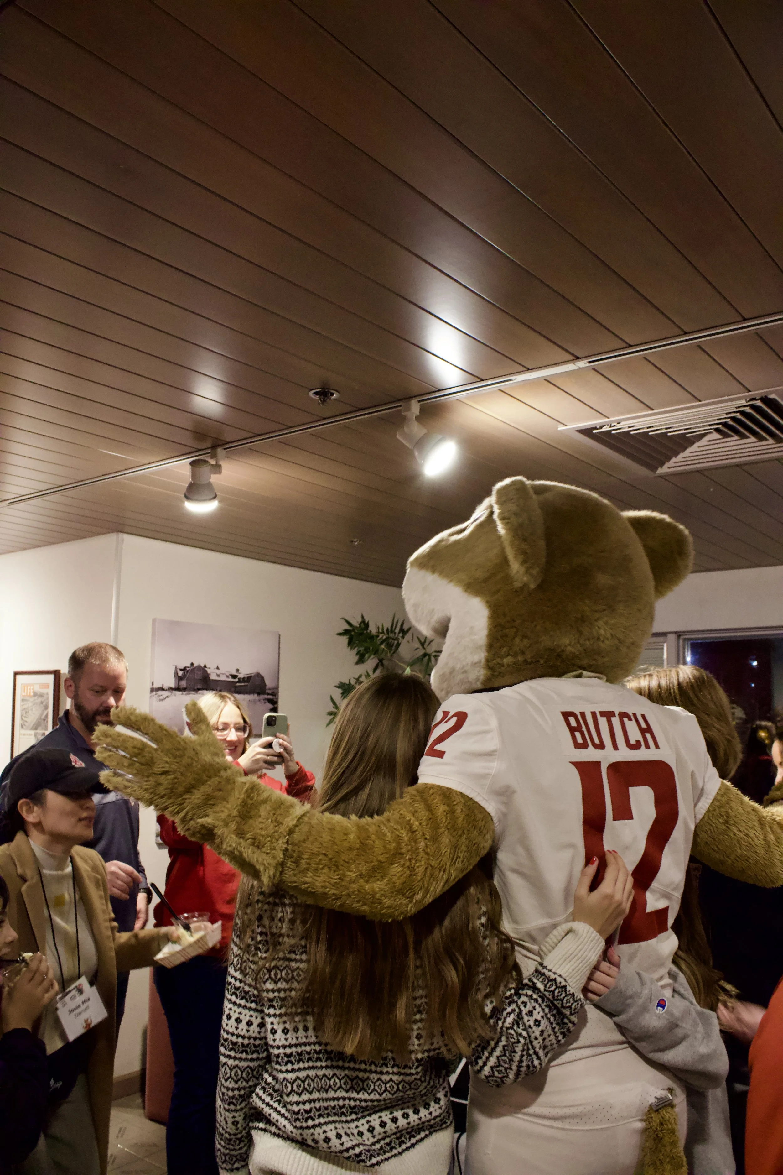 A person in a wolf mascot costume hugs a girl at an indoor gathering, with other people smiling and taking photos in the background.