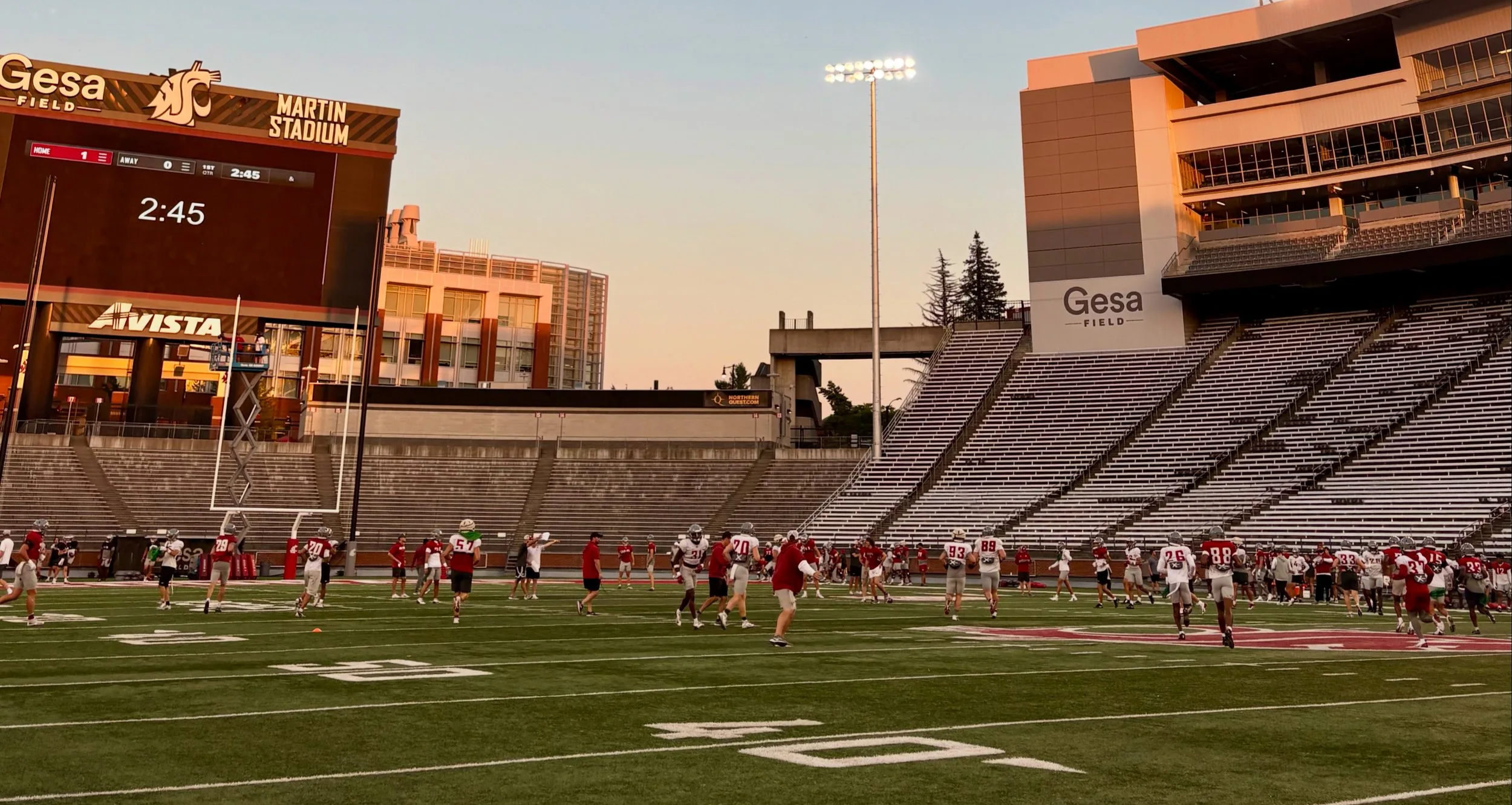 Football players practicing on the field at Gesa Field in the late afternoon, with a large electronic scoreboard showing 2:45, and empty bleachers surrounding the field.